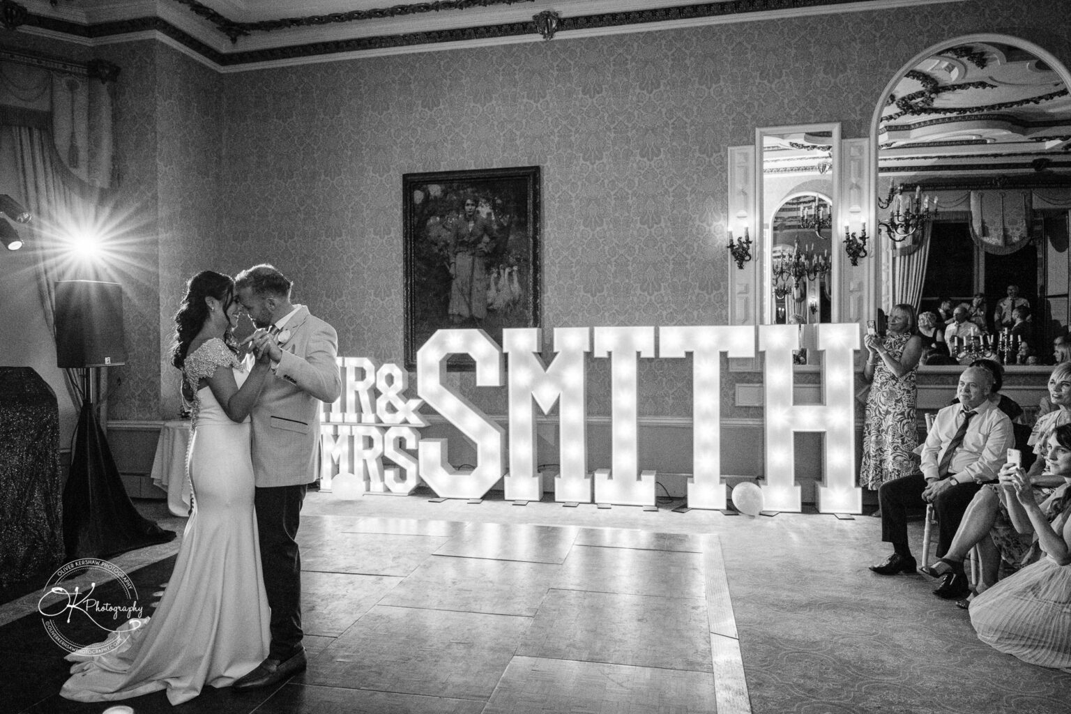 Ettington Park Hotel Wedding Photography A bride and groom share their first dance in an elegant ballroom with large illuminated letters spelling "MR & MRS SMITH" in the background, while guests watch and take photos.