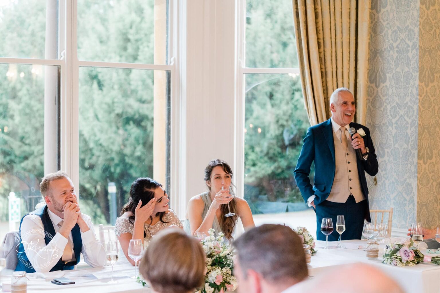 Ettington Park Hotel Wedding Photography An elderly man in a suit giving a speech at a wedding reception, while guests seated at a table listen and smile.