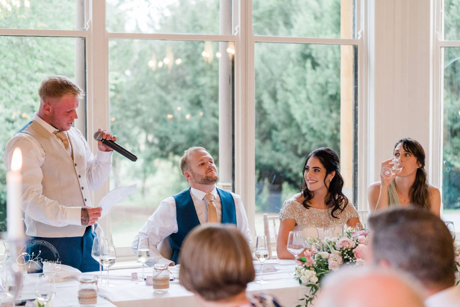 Ettington Park Hotel Wedding Photography A man giving a speech at a wedding reception, while another man, bride, and a woman listen at a table decorated with flowers.