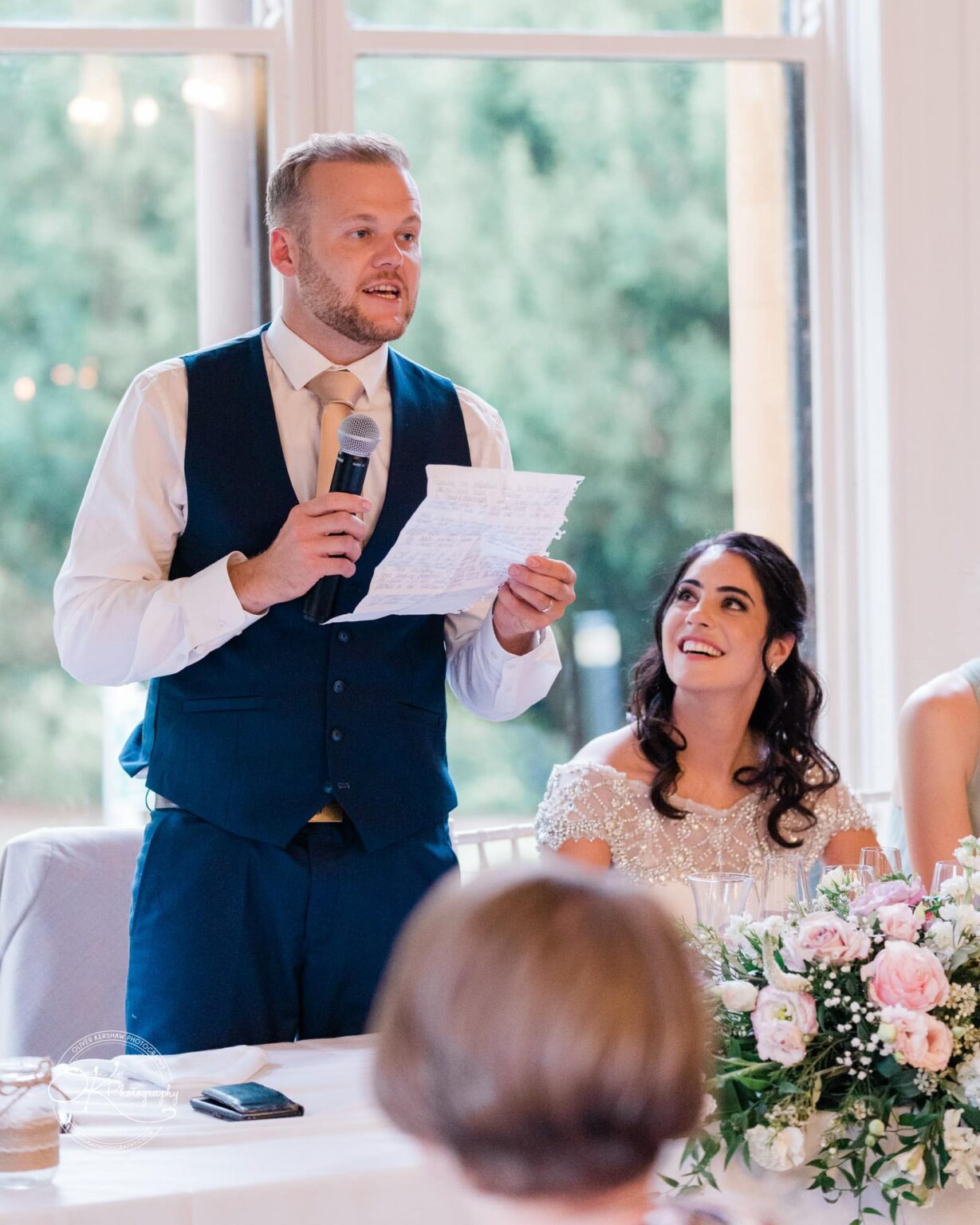 Ettington Park Hotel Wedding Photography A man in a waistcoat and tie gives a speech while holding a microphone and a piece of paper, next to a smiling woman in an off-the-shoulder dress, with a bouquet of flowers on the table.