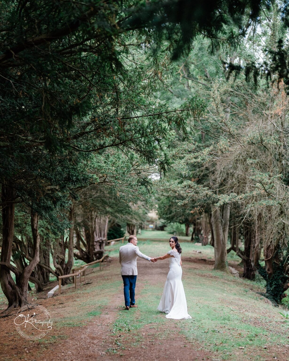 Ettington Park Hotel Wedding Photography A couple holding hands, walking down a tree-lined path, with the bride in a white dress and the groom in a light-coloured suit.