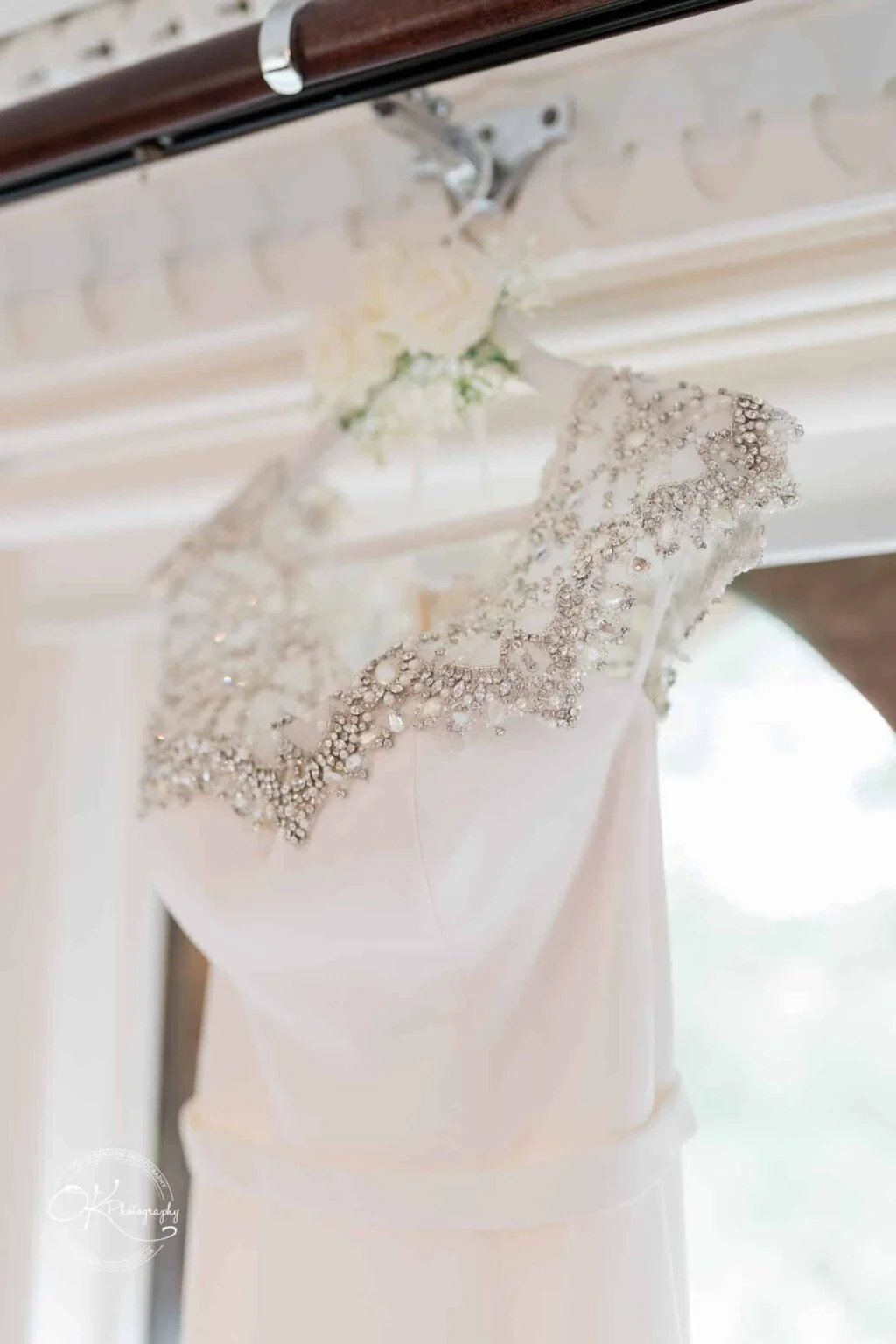 Ettington Park Hotel Wedding Photography A close-up of an elegant white wedding dress with intricate beaded embellishments on the neckline and shoulders, hanging on a decorated hanger.