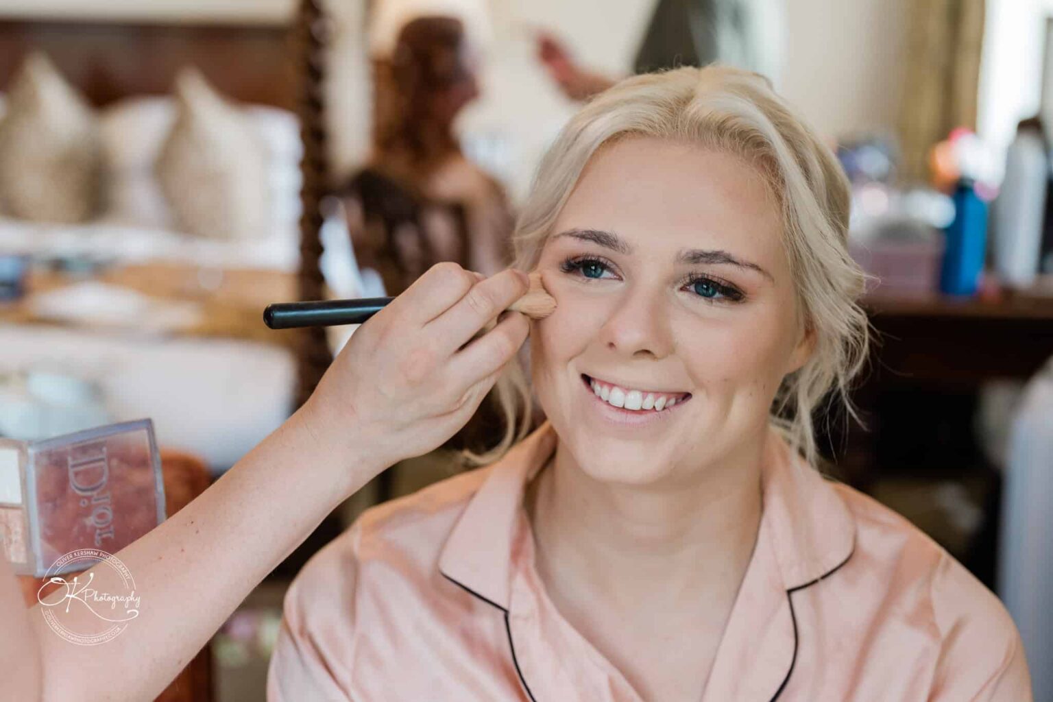 Ettington Park Hotel Wedding Photography A makeup artist applies blush to a smiling blonde woman wearing a pink robe.