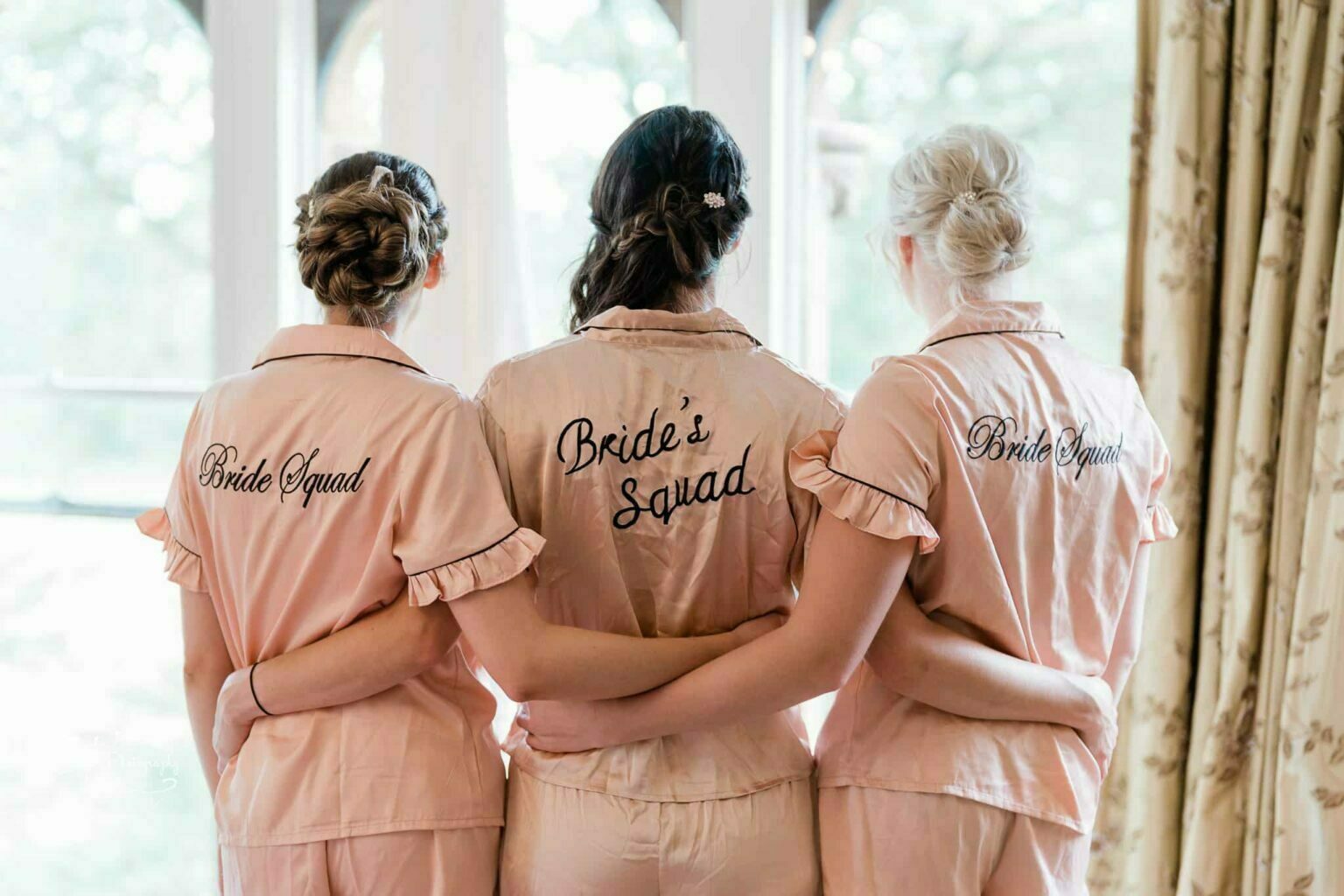 Ettington Park Hotel Wedding Photography Three women in matching pink pyjamas with "Bride Squad" printed on the back, standing with their arms around each other.