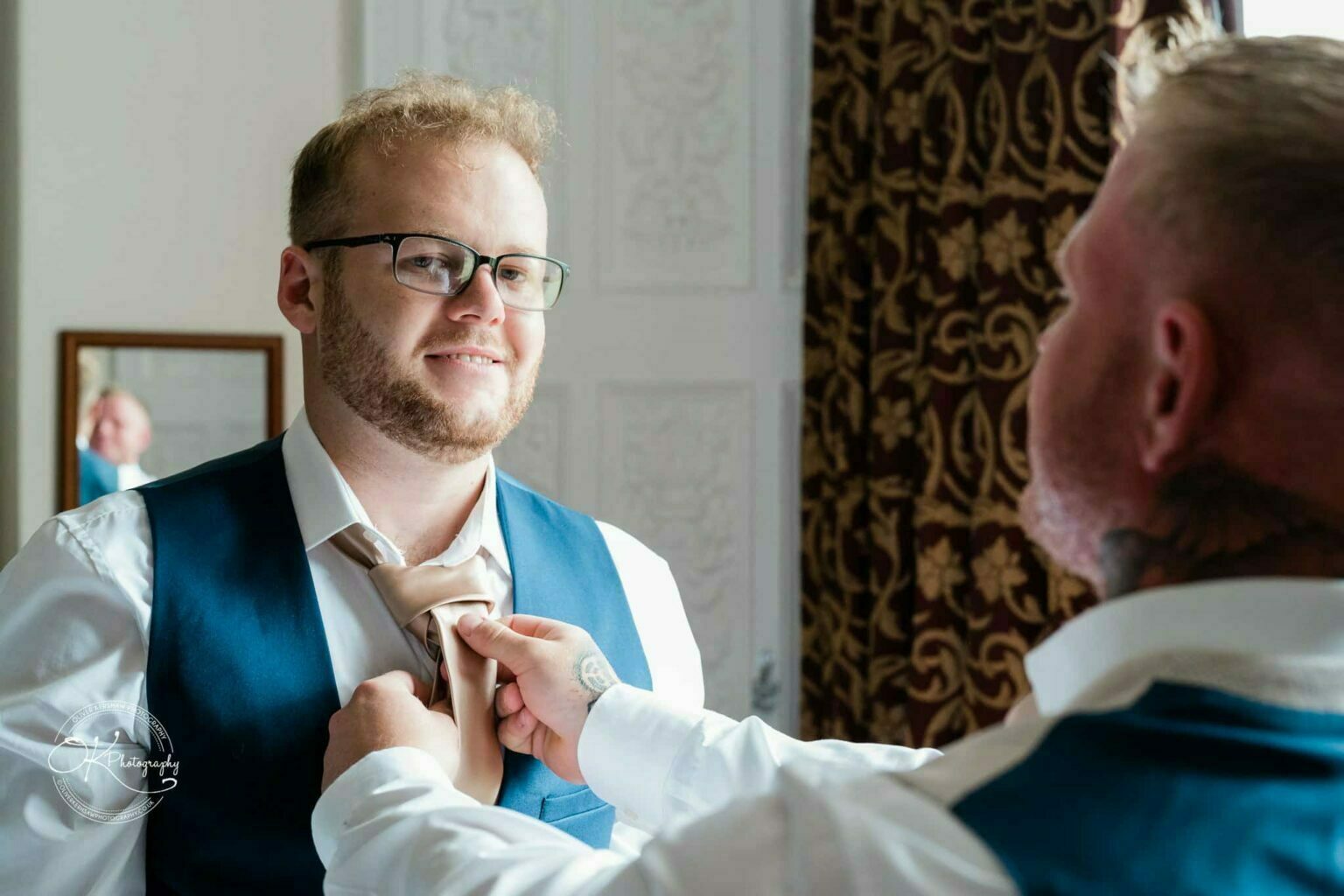 Ettington Park Hotel Wedding Photography A smiling man in glasses and a blue waistcoat has his tie adjusted by another person in preparation for a formal event.
