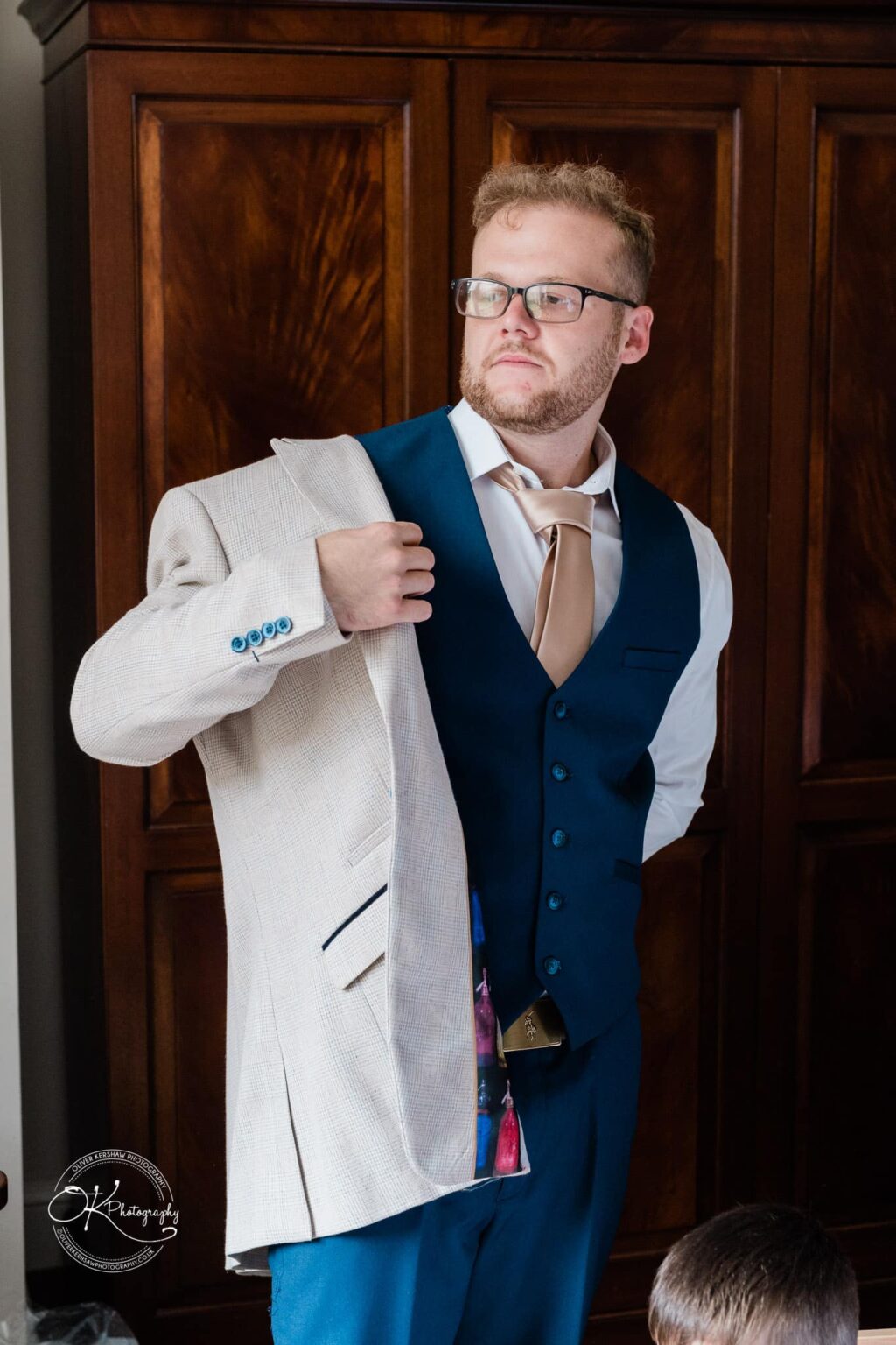 Ettington Park Hotel Wedding Photography Man in a blue waistcoat and beige tie putting on a beige suit jacket, standing in front of a dark wooden cabinet.