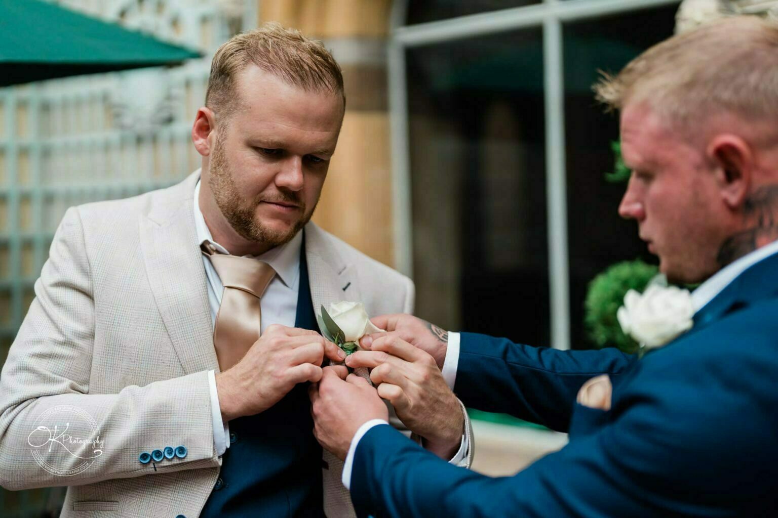 Ettington Park Hotel Wedding Photography A man in a beige suit is having a boutonniere pinned on by another man in a blue suit.