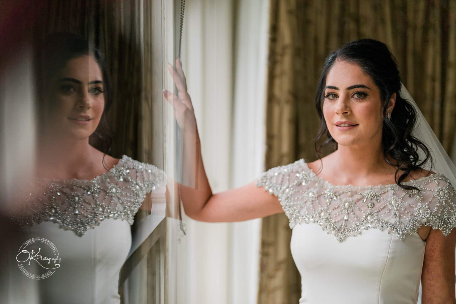 Ettington Park Hotel Wedding Photography Bride in a white wedding dress with beaded embellishments, standing by a window with her reflection visible on the glass.