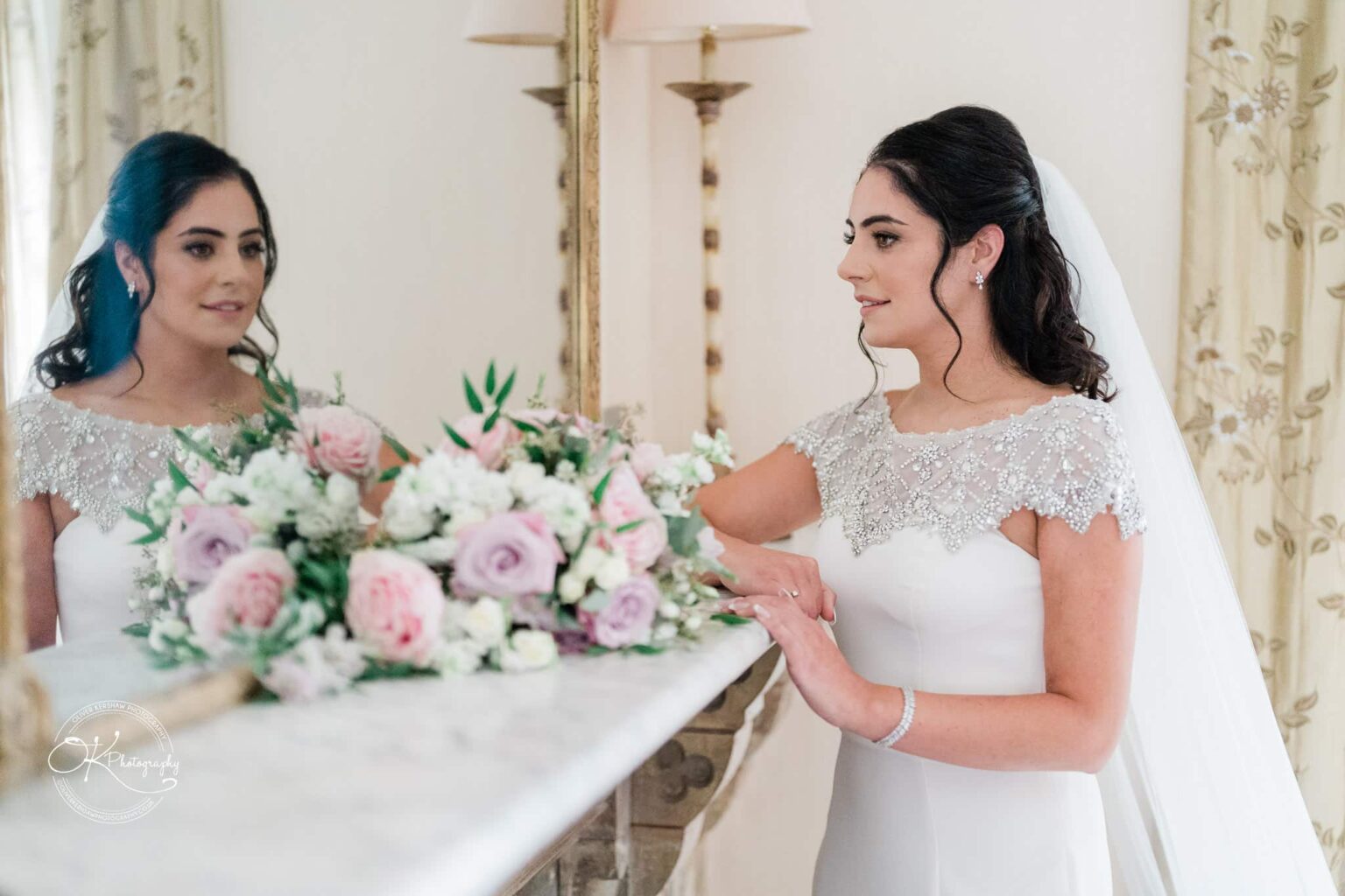 Ettington Park Hotel Wedding Photography A bride in an elegant white dress and veil admires her reflection in a mirror with a bouquet of flowers in front of her.