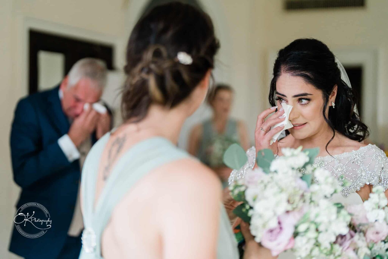 Ettington Park Hotel Wedding Photography Bride wiping a tear from her eye while holding a bouquet, with an older man in the background also wiping his eyes.