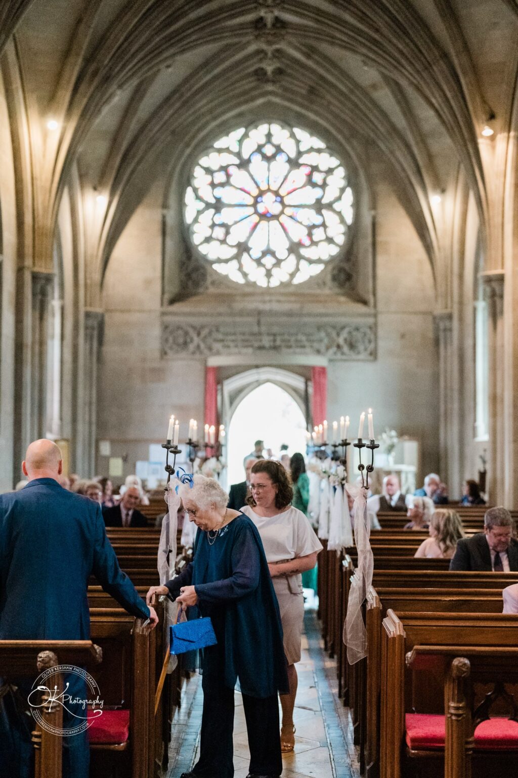 Ettington Park Hotel Wedding Photography People standing and sitting inside a church with a large stained glass window in the background.