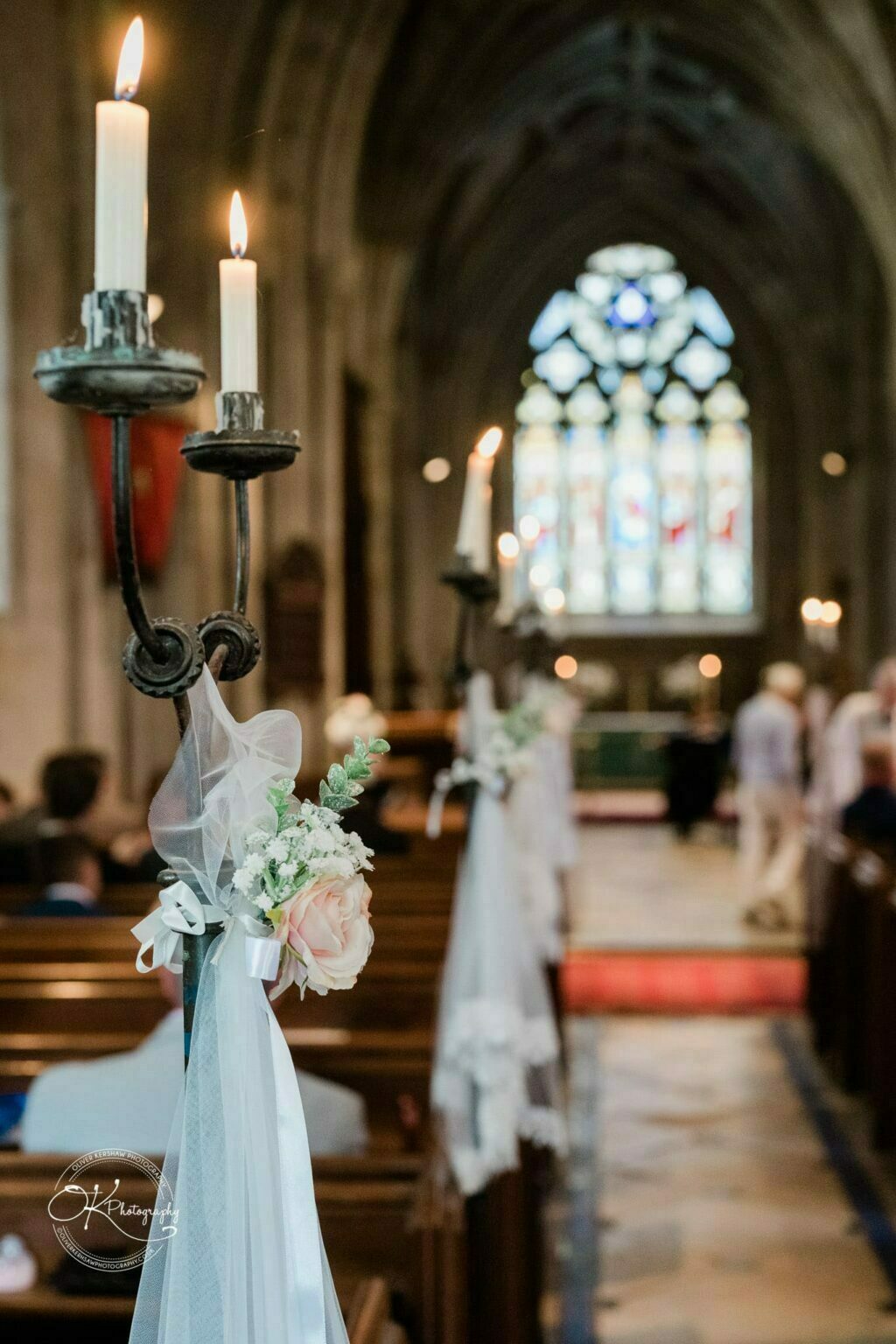Ettington Park Hotel Wedding Photography Lit candles on holders decorated with flowers and white ribbons, lining the aisle of a church with a stained glass window in the background.