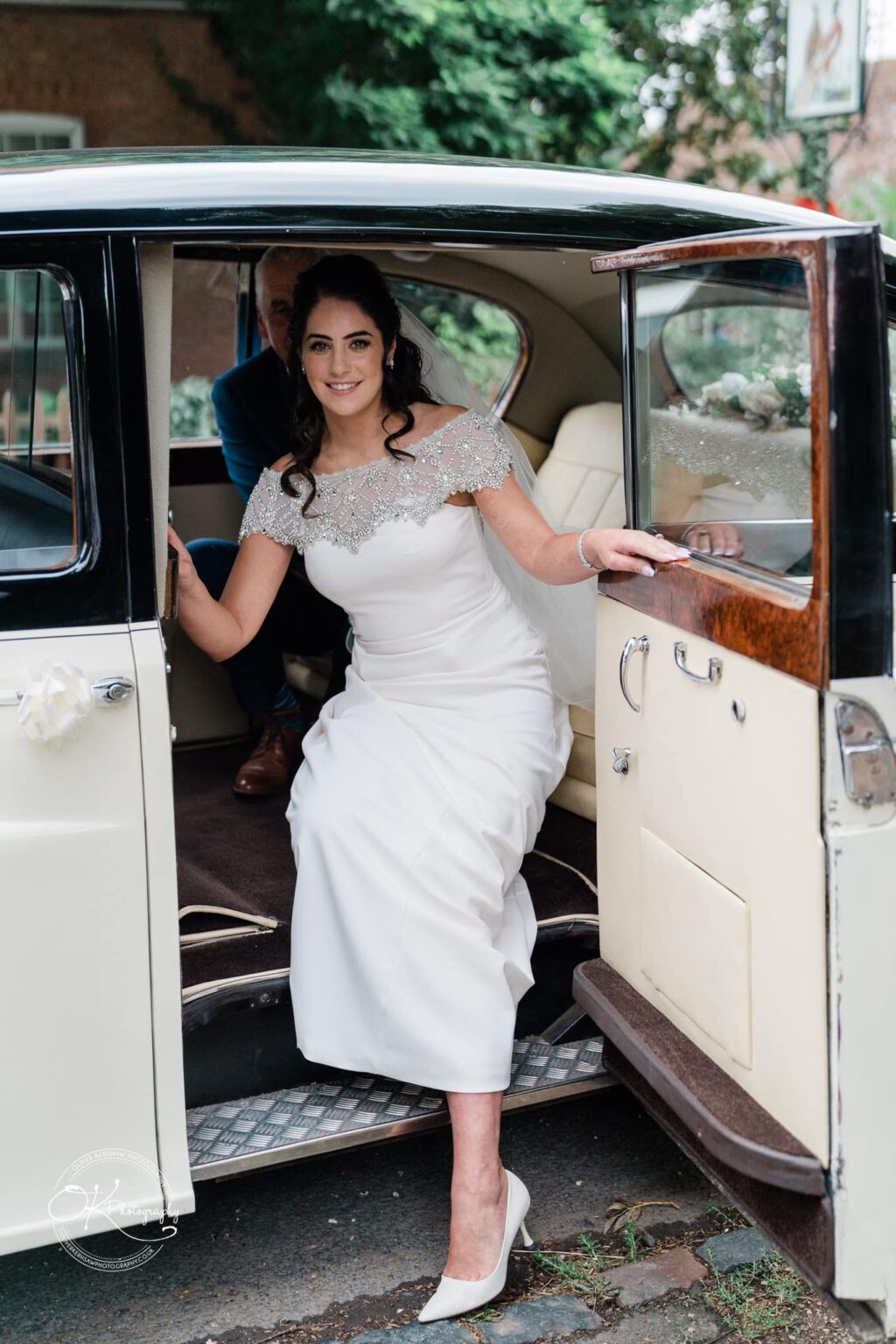 Ettington Park Hotel Wedding Photography Bride stepping out of a vintage car in a white wedding dress, smiling towards the camera.
