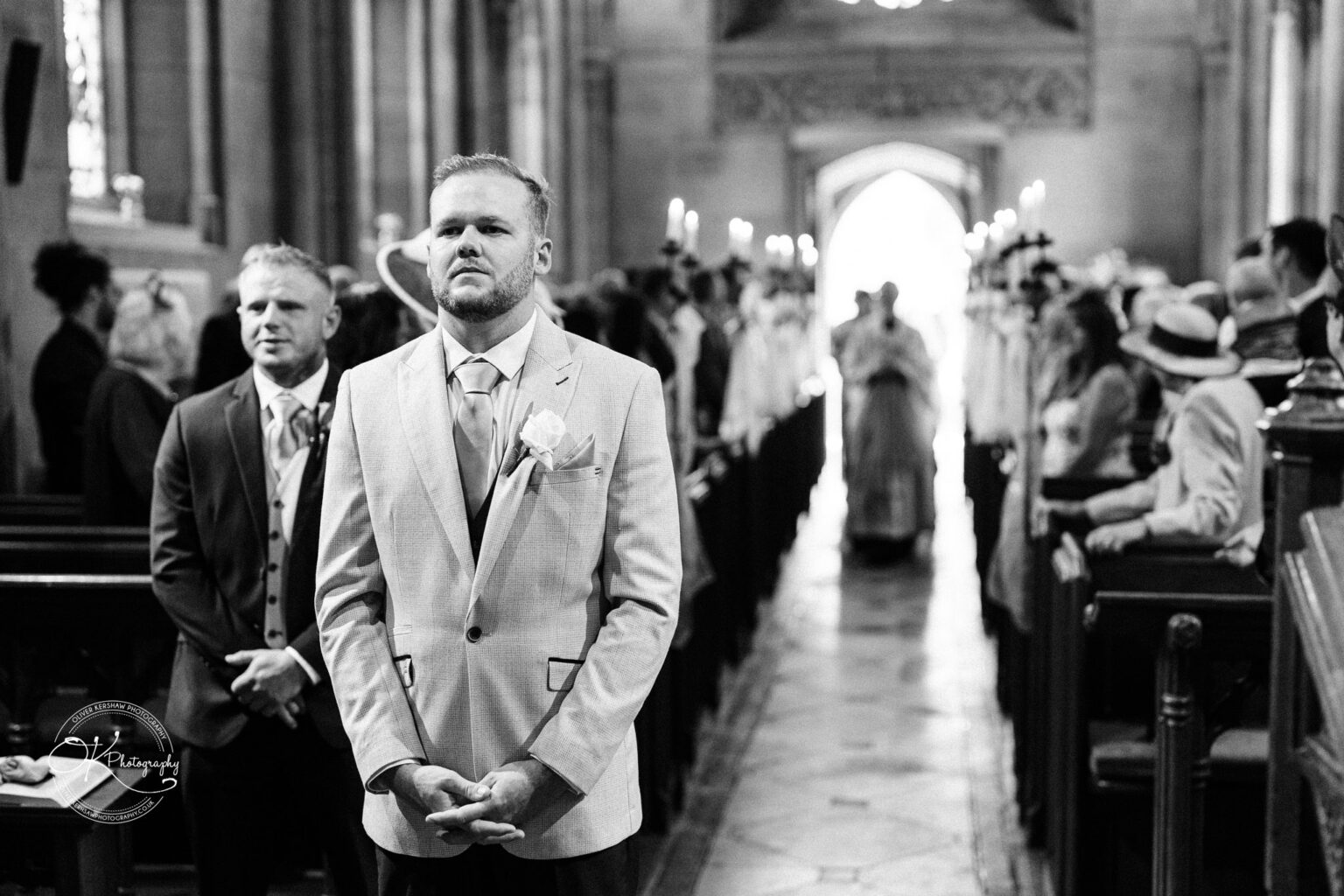 Ettington Park Hotel Wedding Photography Groom anxiously waiting at the altar of a church, with guests and a priest approaching in the background.
