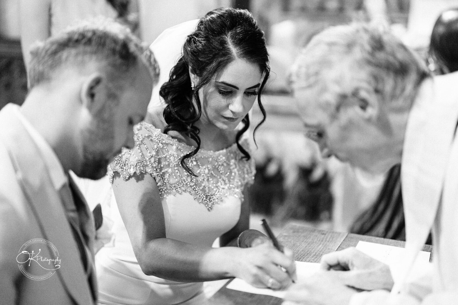 Ettington Park Hotel Wedding Photography A bride signs a document while sits a groom beside her and an officiant leans over a table.