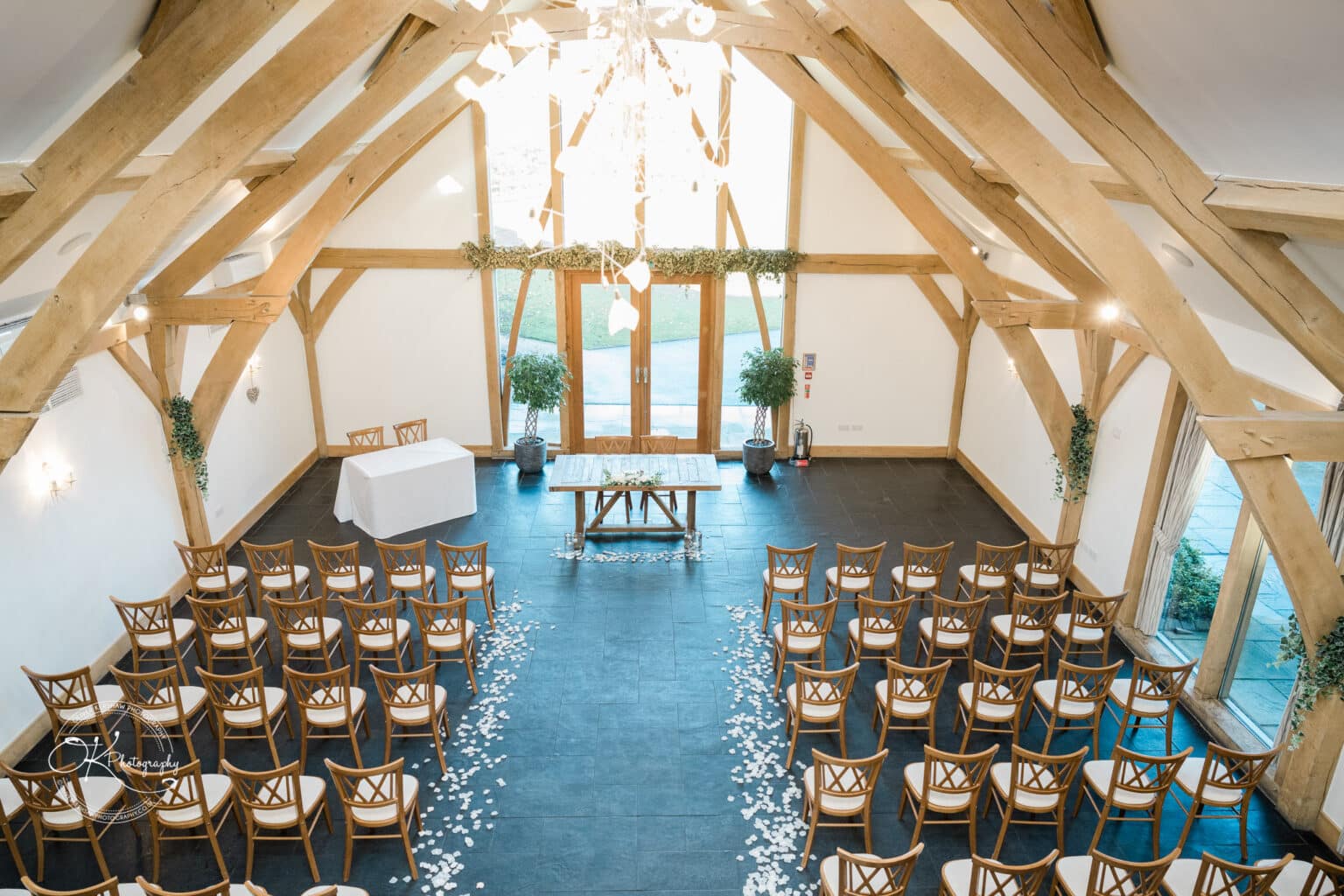 Mythe Barn Wedding photography Aerial view of an indoor wedding ceremony setup at Mythe Barn with wooden chairs arranged neatly, a flower petal aisle, a signing table, and lush greenery decor.