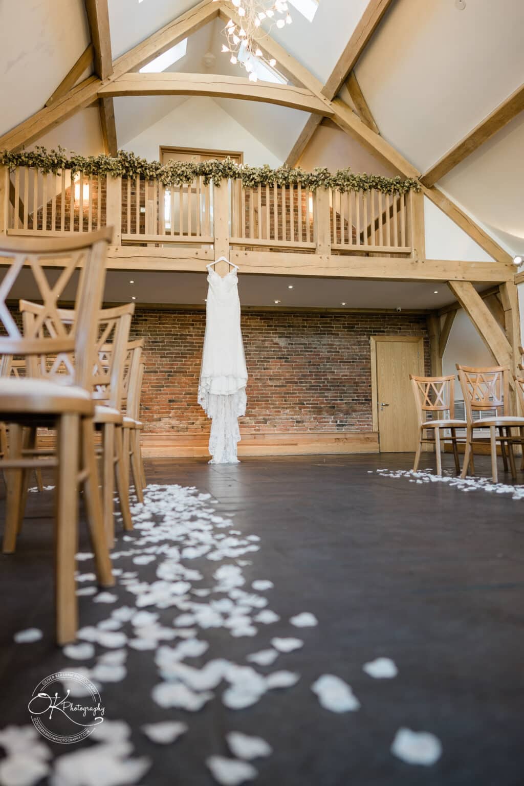 Mythe Barn Wedding photography A wedding dress hanging from a wooden balcony inside a barn decorated with greenery, with white flower petals scattered on the floor and wooden chairs set up on either side.