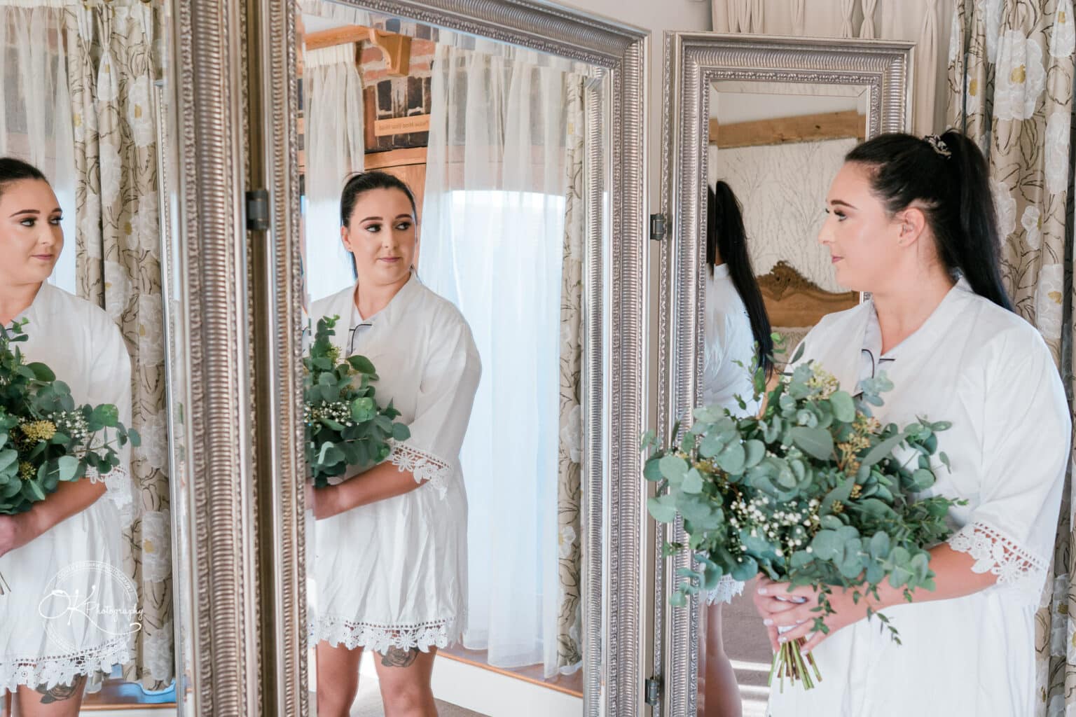 Mythe Barn Wedding photography Bride in a white robe holding a green bouquet, standing in front of a large mirror reflecting her image, with floral curtains and rustic decor in the background.