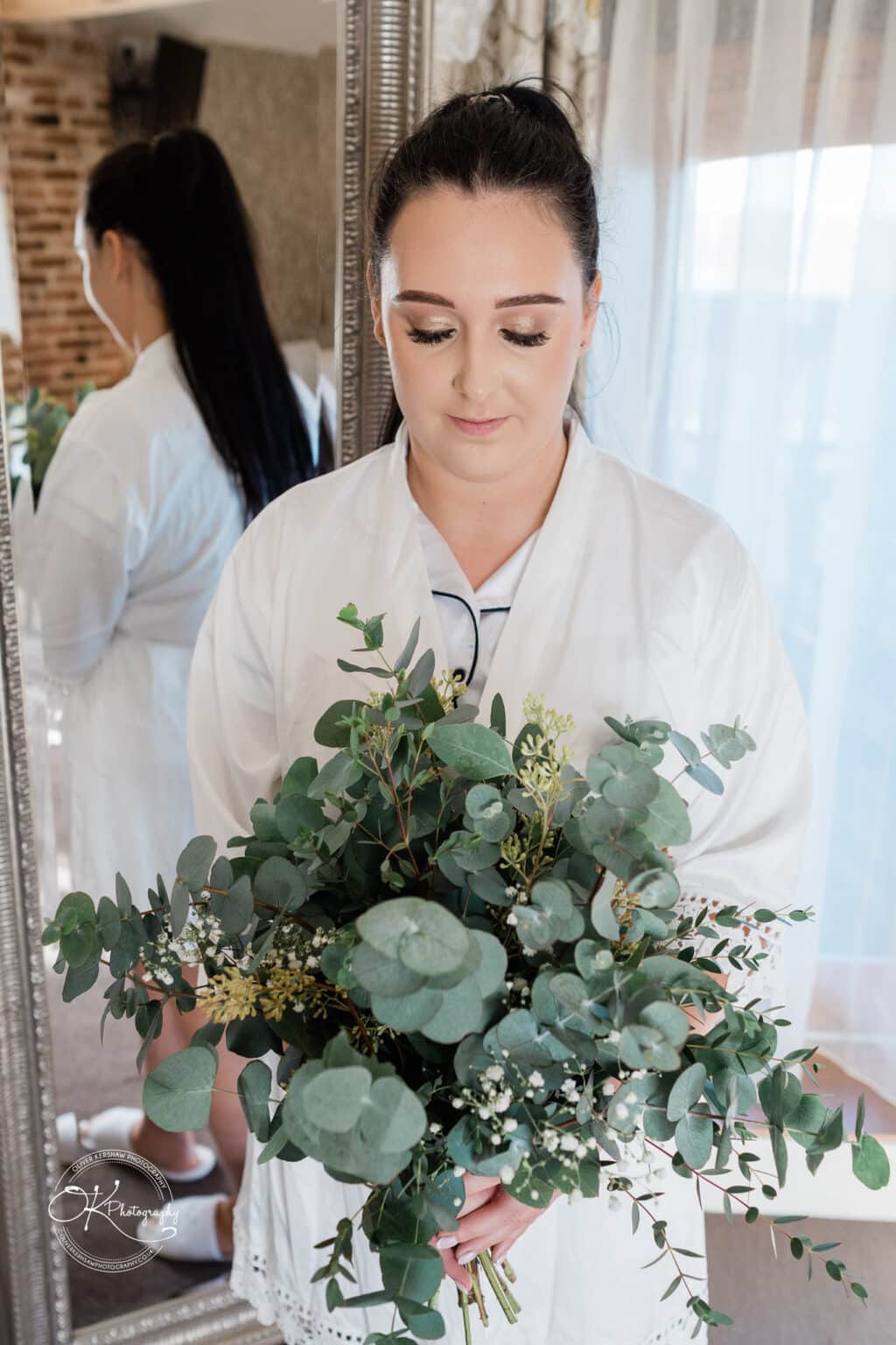 Mythe Barn Wedding photography Bride holding a bouquet of greenery and white flowers, standing in front of a mirror at Mythe Barn.