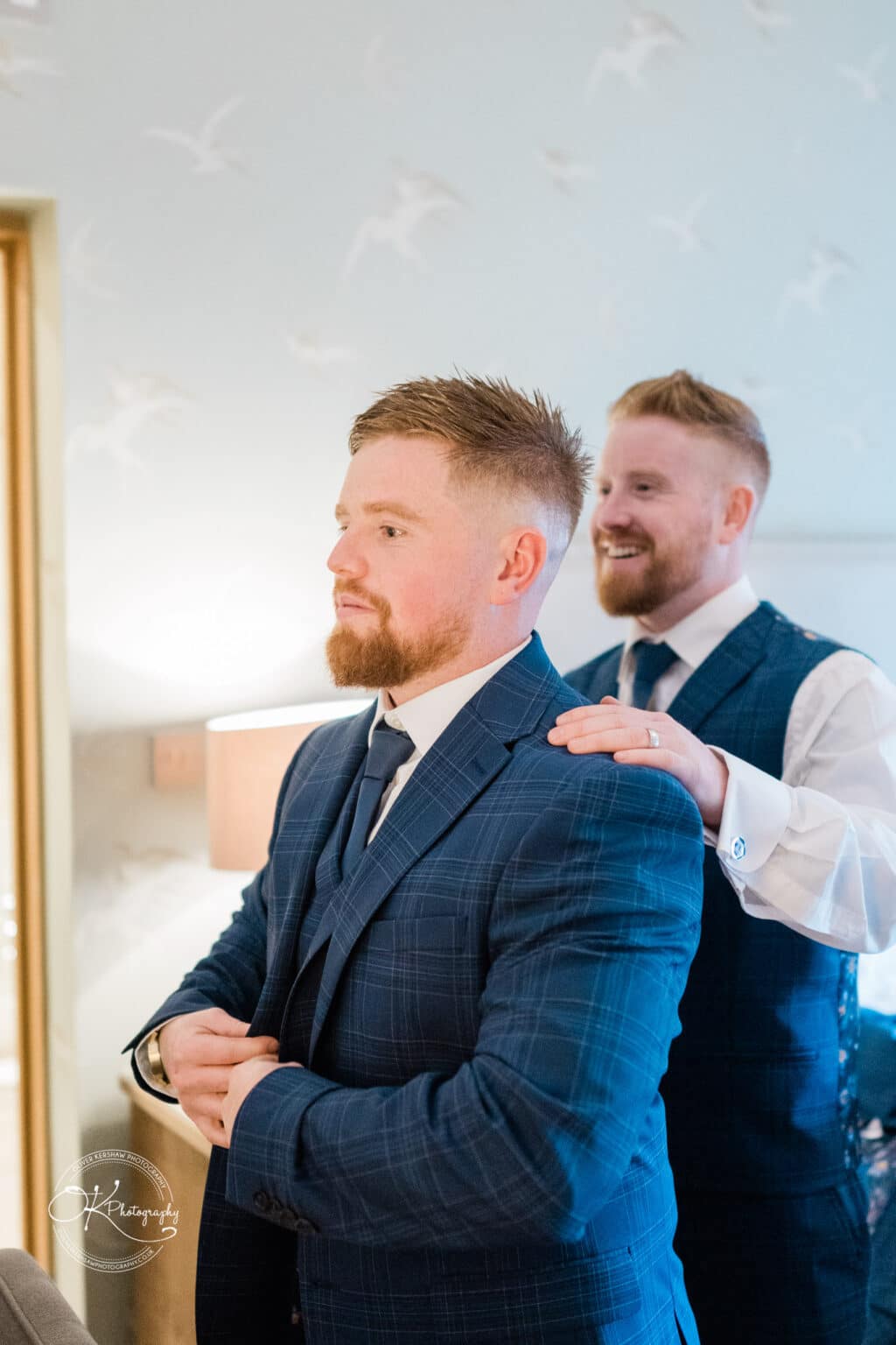 Mythe Barn Wedding photography Two groomsmen in blue checkered suits, with one assisting the other in fastening his jacket.