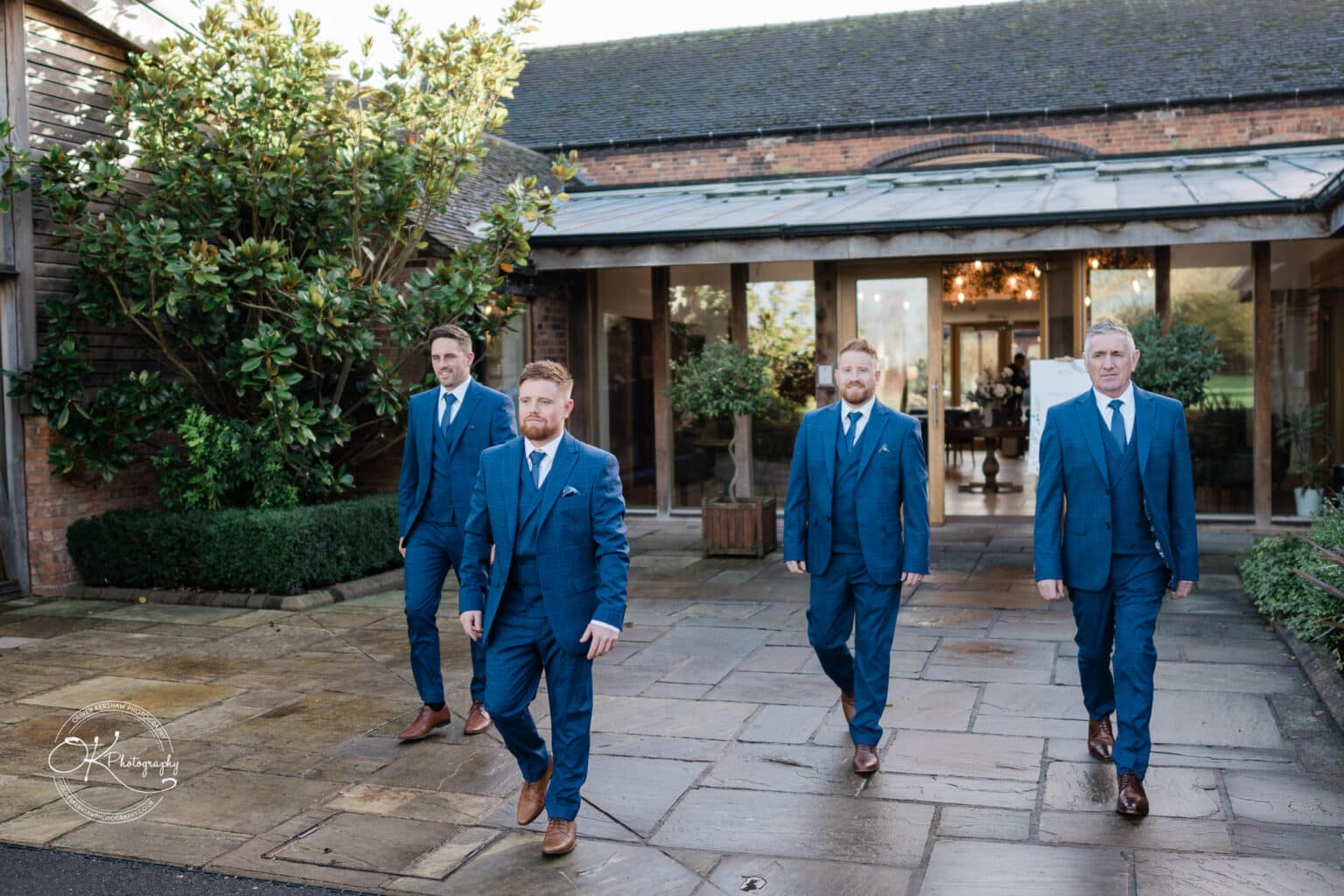 Mythe Barn Wedding photography Four men in blue suits walking outside a rustic barn with glass doors and a large bush in the background.