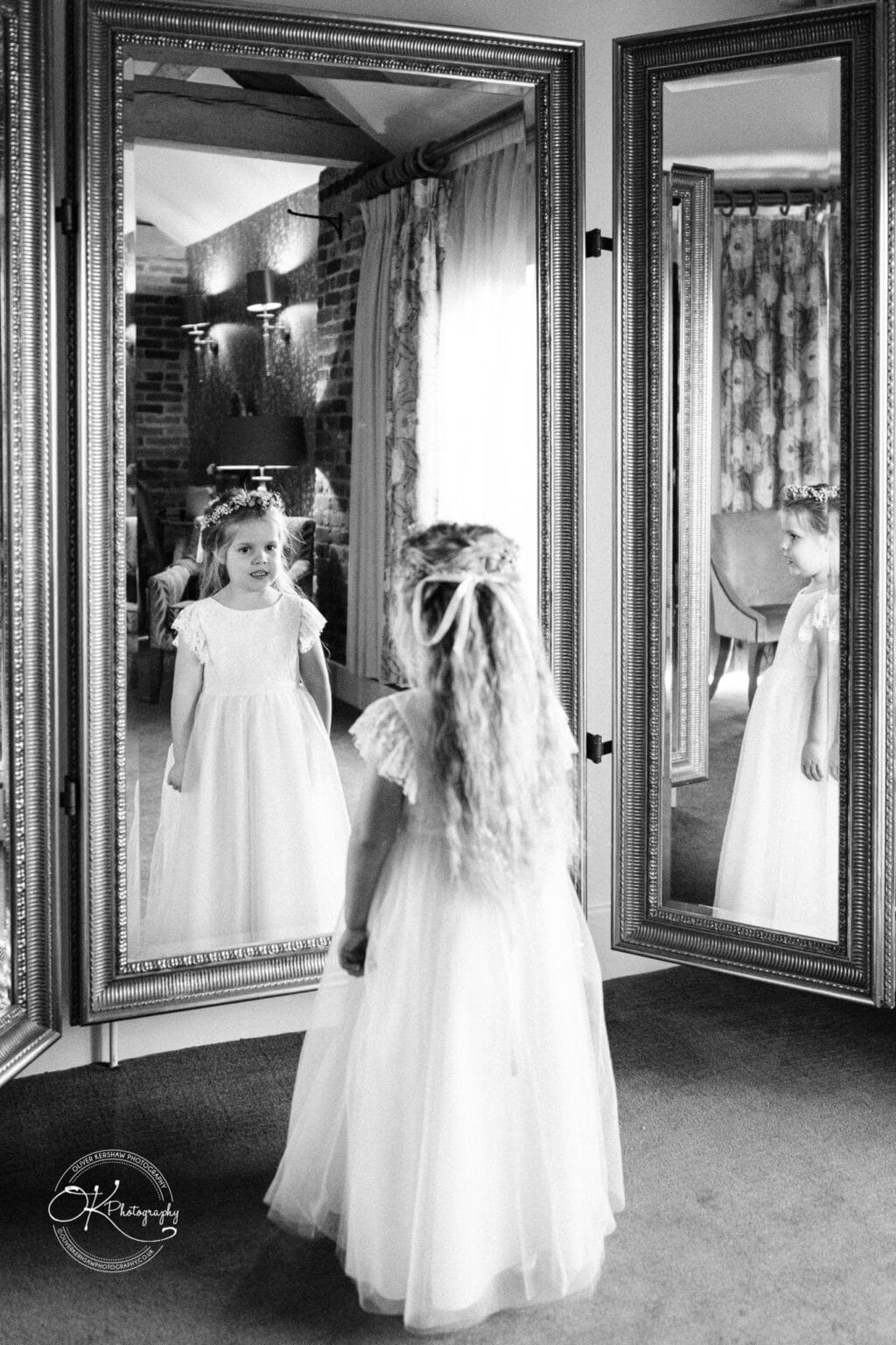 Mythe Barn Wedding photography A young girl in a white dress and flower crown looks at her reflection in a large ornate mirror.