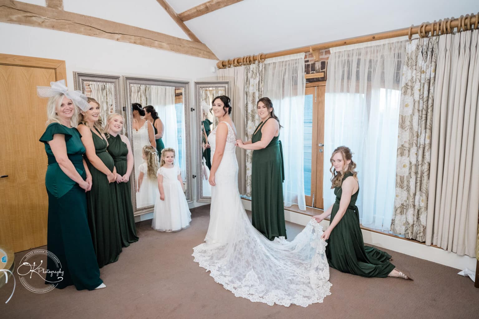 Mythe Barn Wedding photography Bride in wedding dress surrounded by bridesmaids in green dresses and a flower girl in white, preparing in a rustic room with exposed wooden beams and mirrored wardrobe doors.