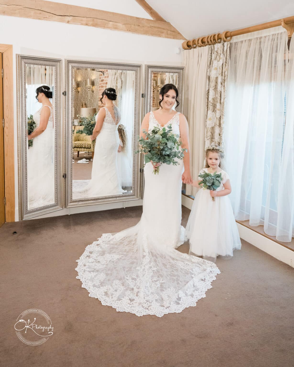 Mythe Barn Wedding photography Bride holding a bouquet, standing with a young flower girl in a room with large mirrors and lace curtains.