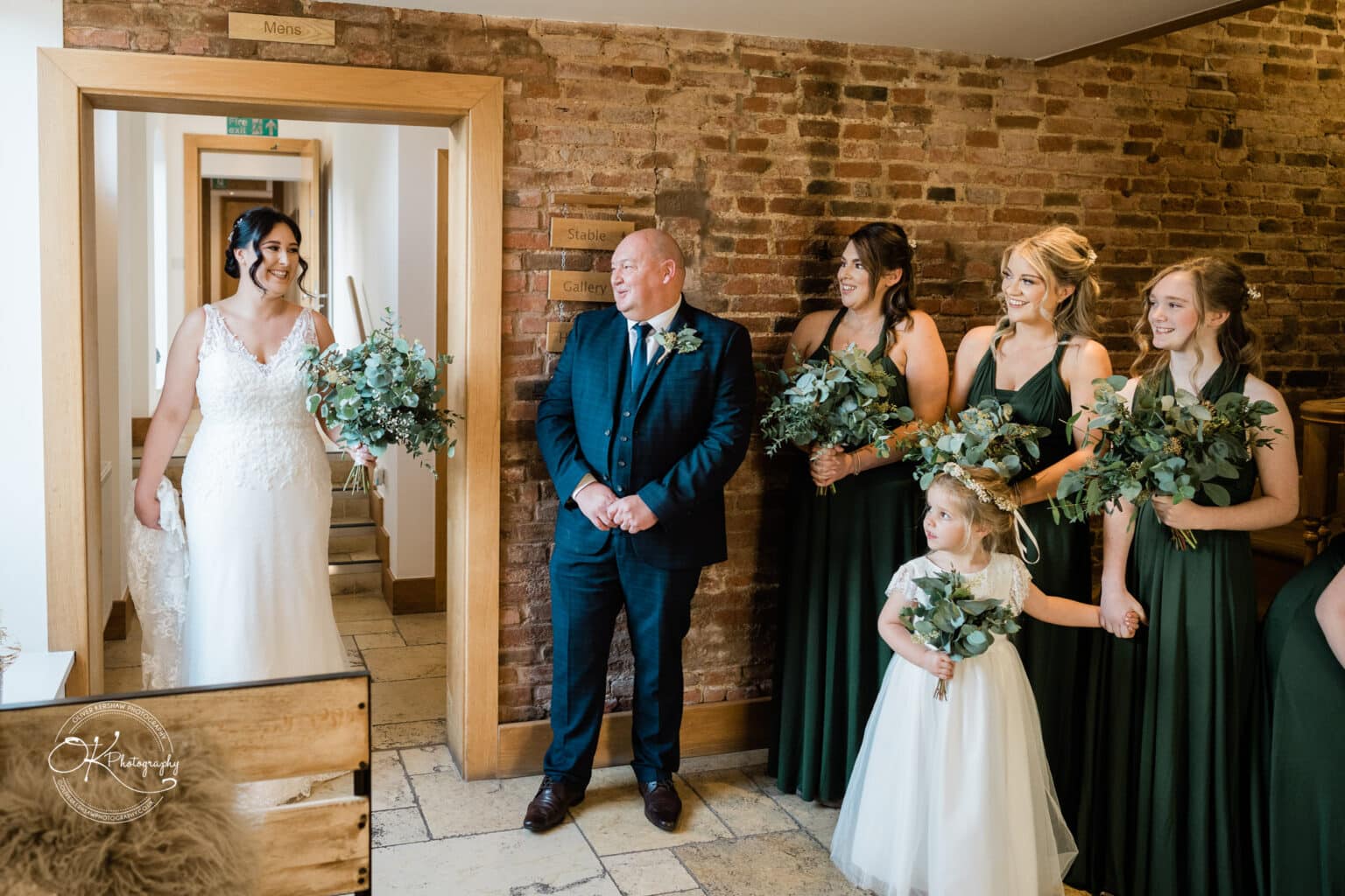 Mythe Barn Wedding photography Bride in a white gown holding a bouquet, entering a room with a man and bridesmaids in dark green dresses, all smiling and holding bouquets at Mythe Barn wedding venue.