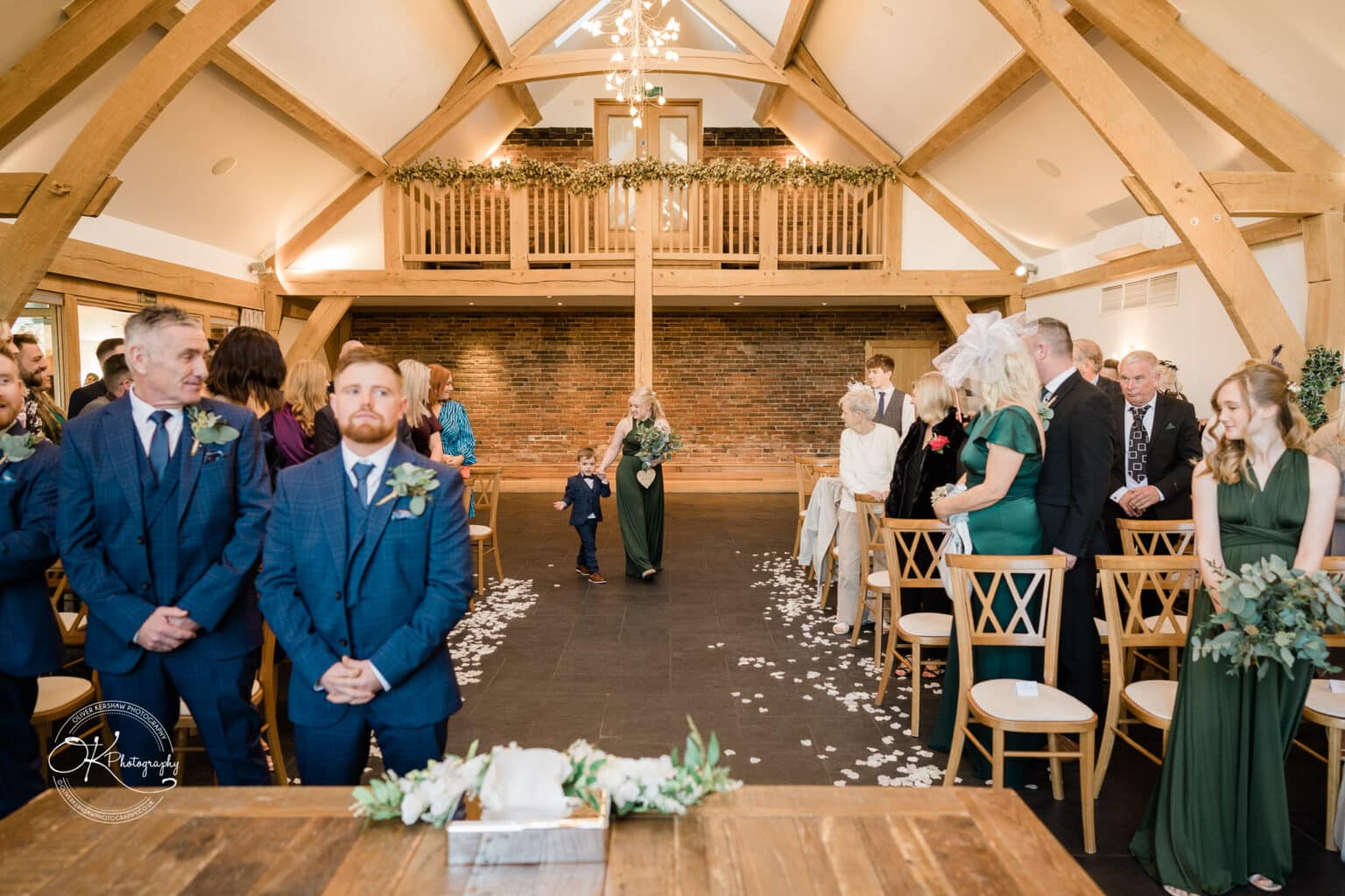 Mythe Barn Wedding photography A wedding ceremony at Mythe Barn showcasing an aisle decorated with white petals, guests in semi-formal attire, wooden beams, and rustic decor.