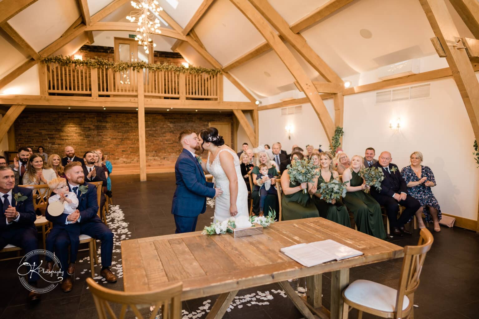Mythe Barn Wedding photography A bride and groom kissing during their ceremony in a barn-style venue with wooden beams, surrounded by guests who are clapping and smiling.
