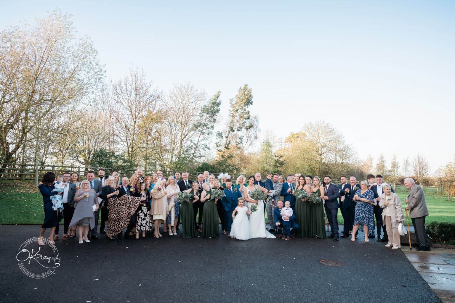 Mythe Barn Wedding photography A large wedding group posing outdoors with trees and clear sky in the background.