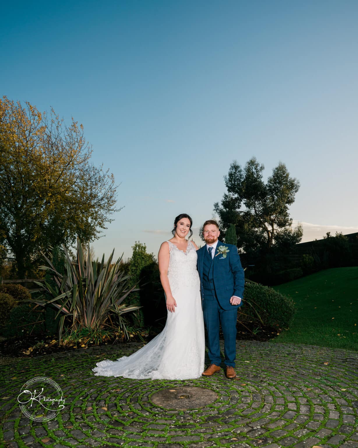 Mythe Barn Wedding photography A bride and groom stand outside on a circular cobblestone path surrounded by green foliage, posing for their wedding photo.