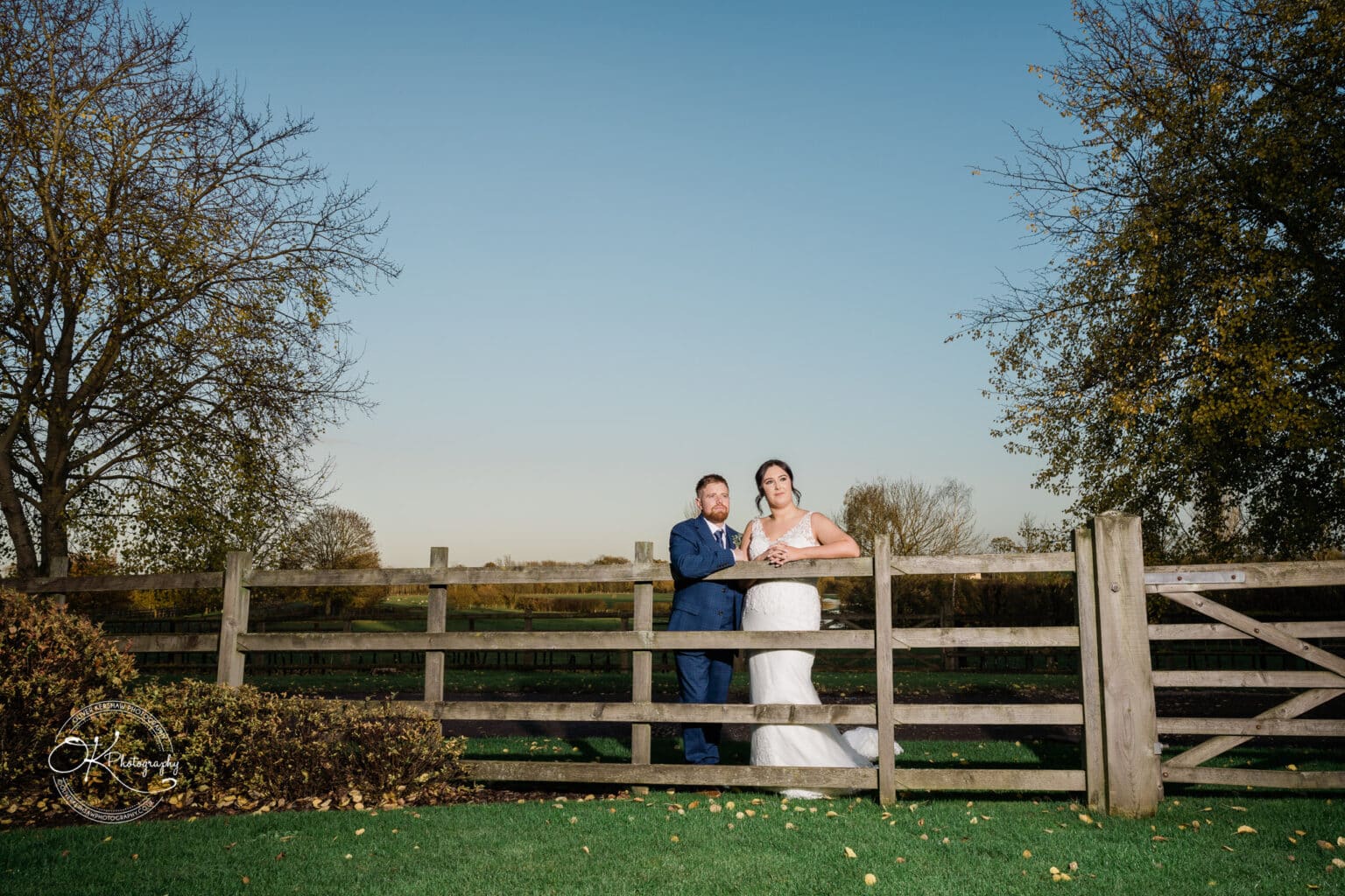 Mythe Barn Wedding photography Bride and groom leaning on a wooden fence in a countryside setting, with trees and a clear sky in the background.