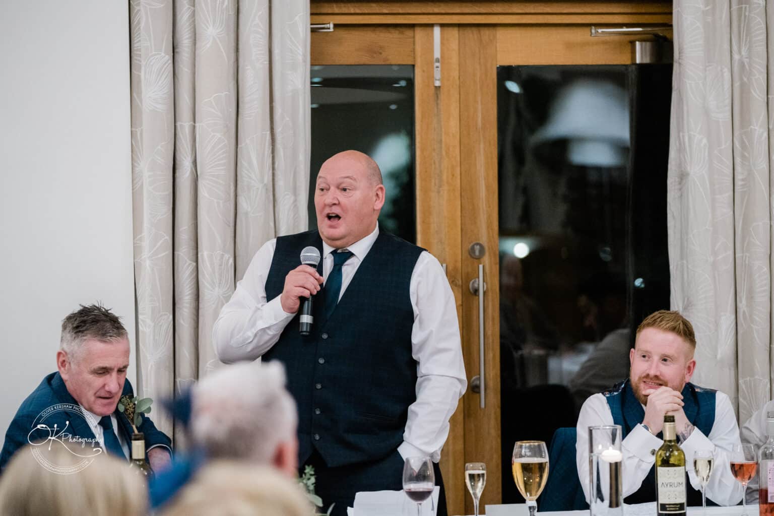 Mythe Barn Wedding photography A man in a waistcoat giving a speech at a wedding reception while holding a microphone, with two other men seated beside him at a table with drinks.