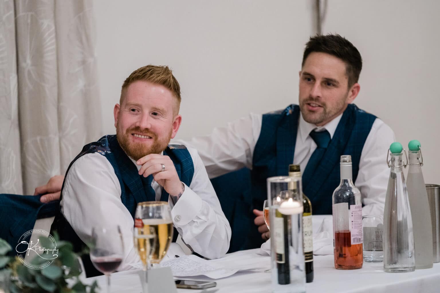 Mythe Barn Wedding photography Two men in suits sit at a table with bottles, glasses, and a candle, enjoying a moment at a wedding reception.