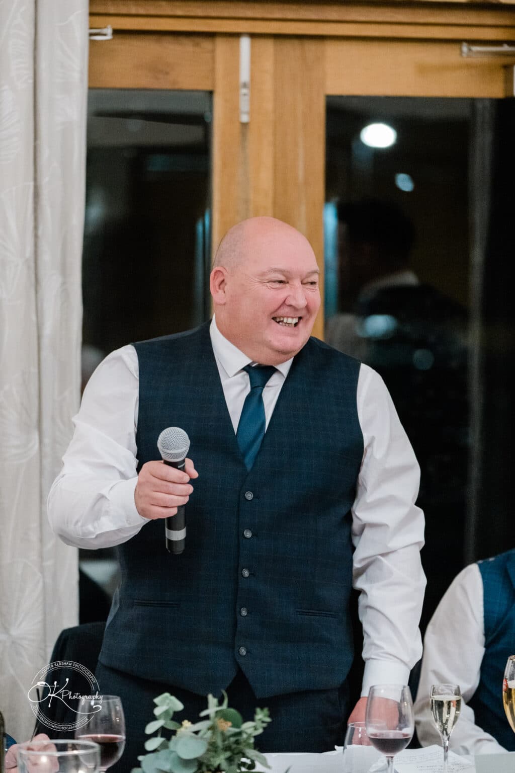 Mythe Barn Wedding photography A man in a dark blue waistcoat and tie, holding a microphone and smiling during a speech at a wedding reception.