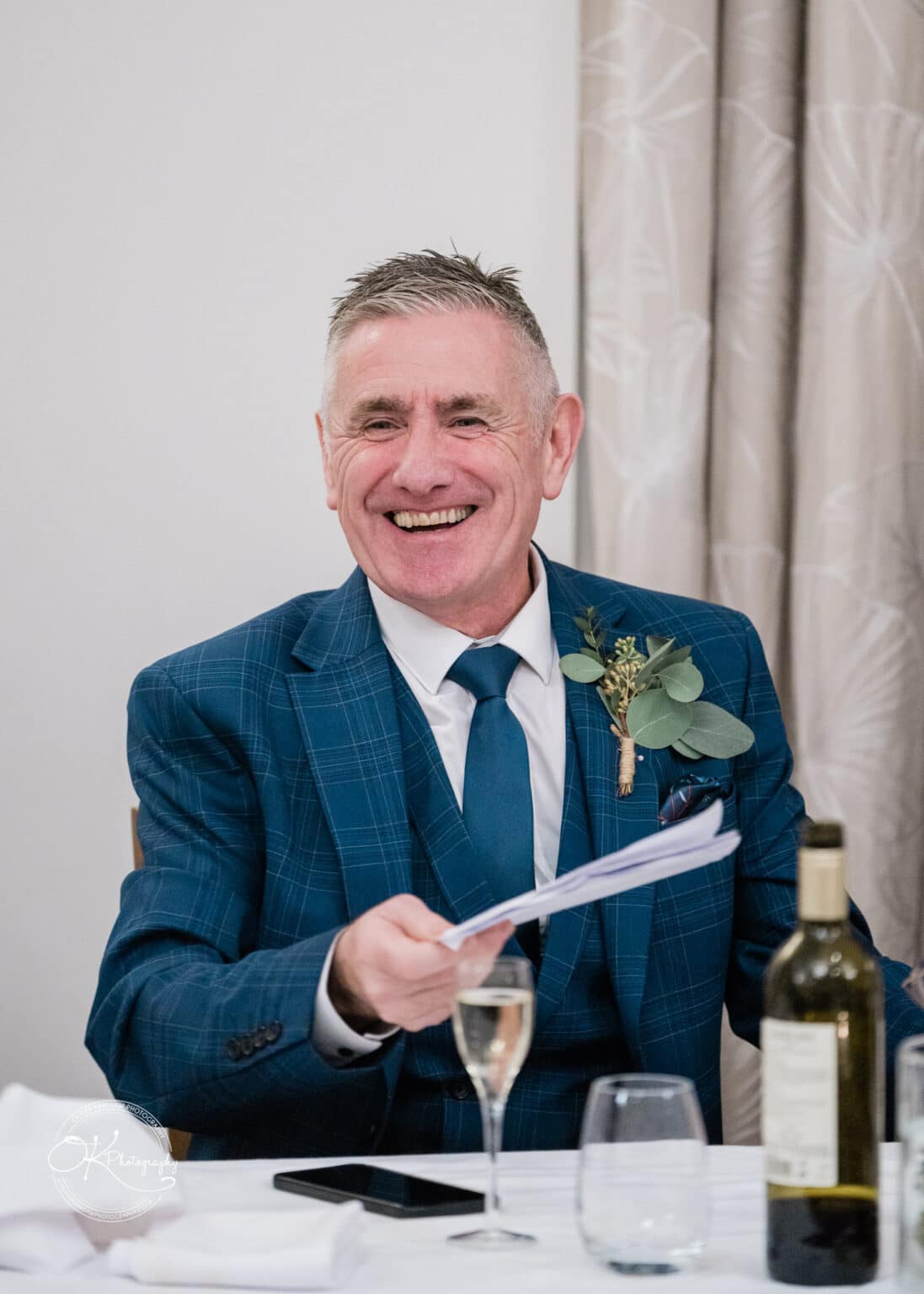 Mythe Barn Wedding photography A man in a blue checkered suit, holding a piece of paper and smiling, sits at a table with a glass of champagne and a bottle of wine.