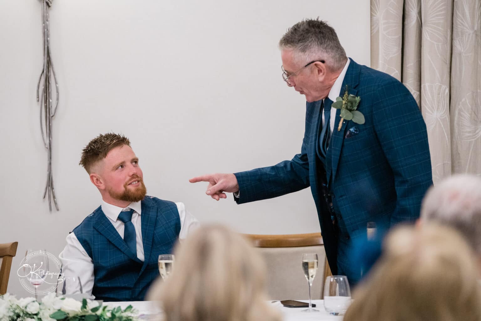 Mythe Barn Wedding photography A wedding speech moment with a seated man looking up at an older man pointing at him, both wearing matching blue suits, in a decorated venue with champagne glasses on the table.