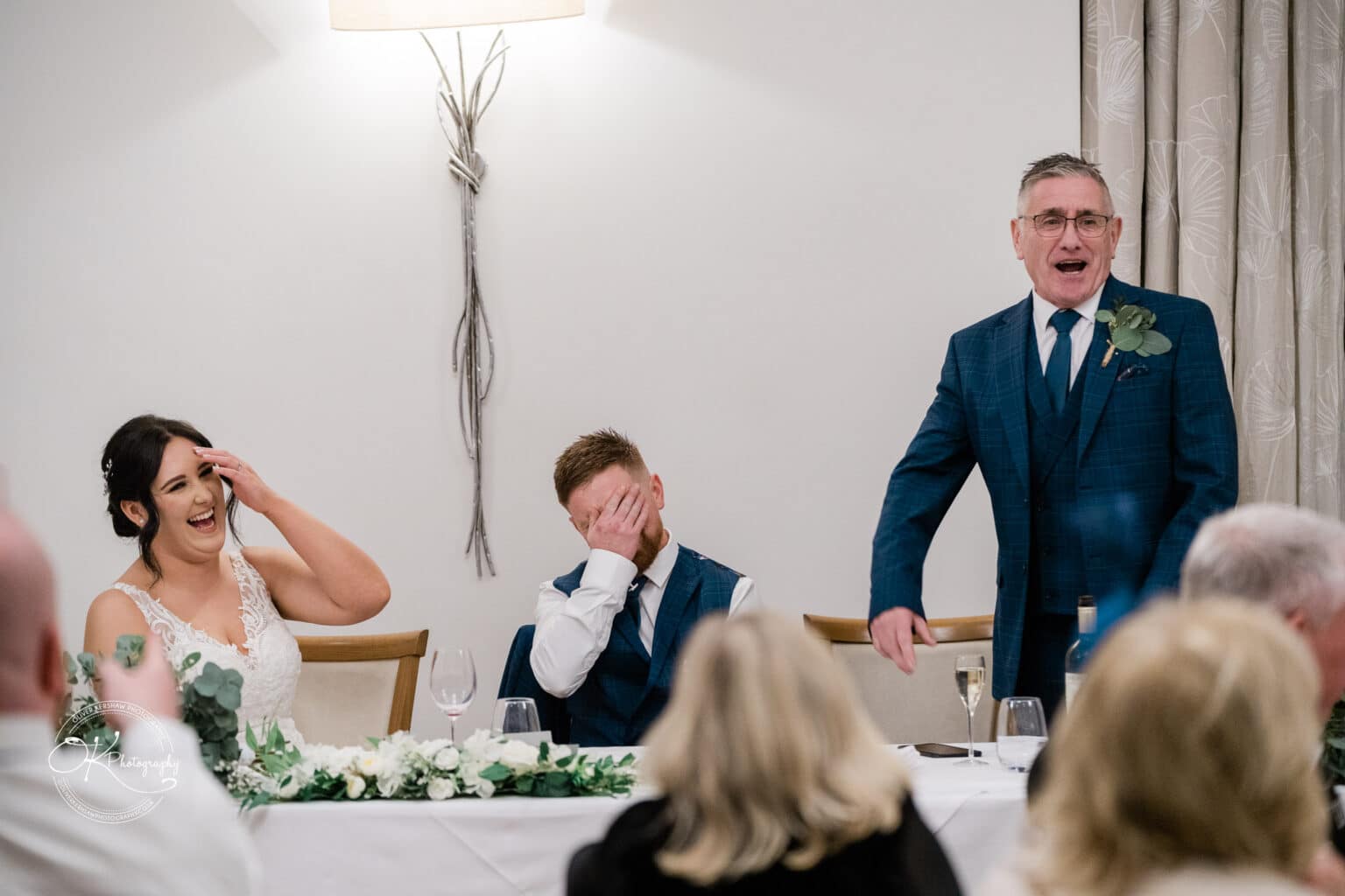 Mythe Barn Wedding photography People laughing and covering faces during a wedding speech at Mythe Barn, with a bride in a white dress, a groom in a blue suit, and a speaker also in a blue suit.