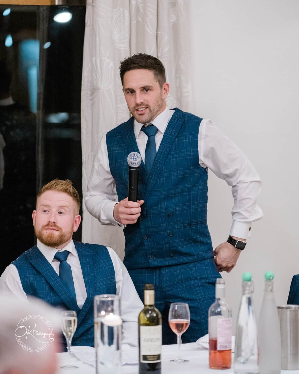 Mythe Barn Wedding photography Two men in blue checkered waistcoats and ties, one standing and holding a microphone while the other sits at a table with drinks and glass bottles.