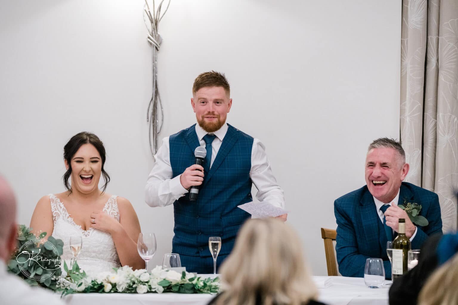 Mythe Barn Wedding photography People laughing while a man in a blue suit speaks into a microphone at a wedding reception.