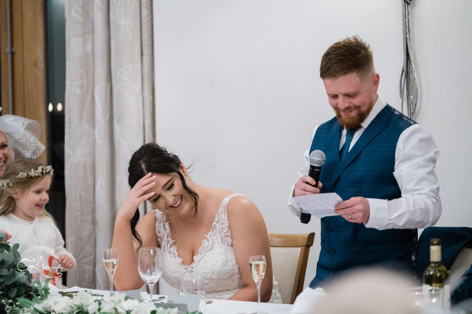 Mythe Barn Wedding photography Man in a blue waistcoat giving a wedding speech while the bride laughs and hides her face with her hand.