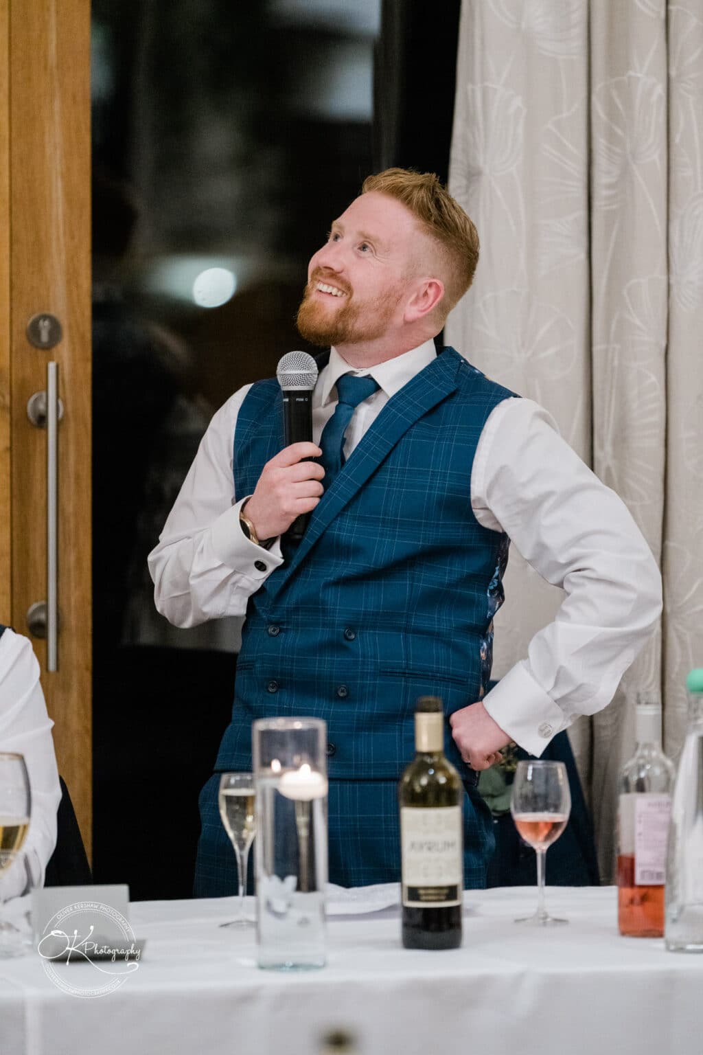 Mythe Barn Wedding photography Man in a blue checkered waistcoat and tie holding a microphone, standing by a table with wine and water bottles.