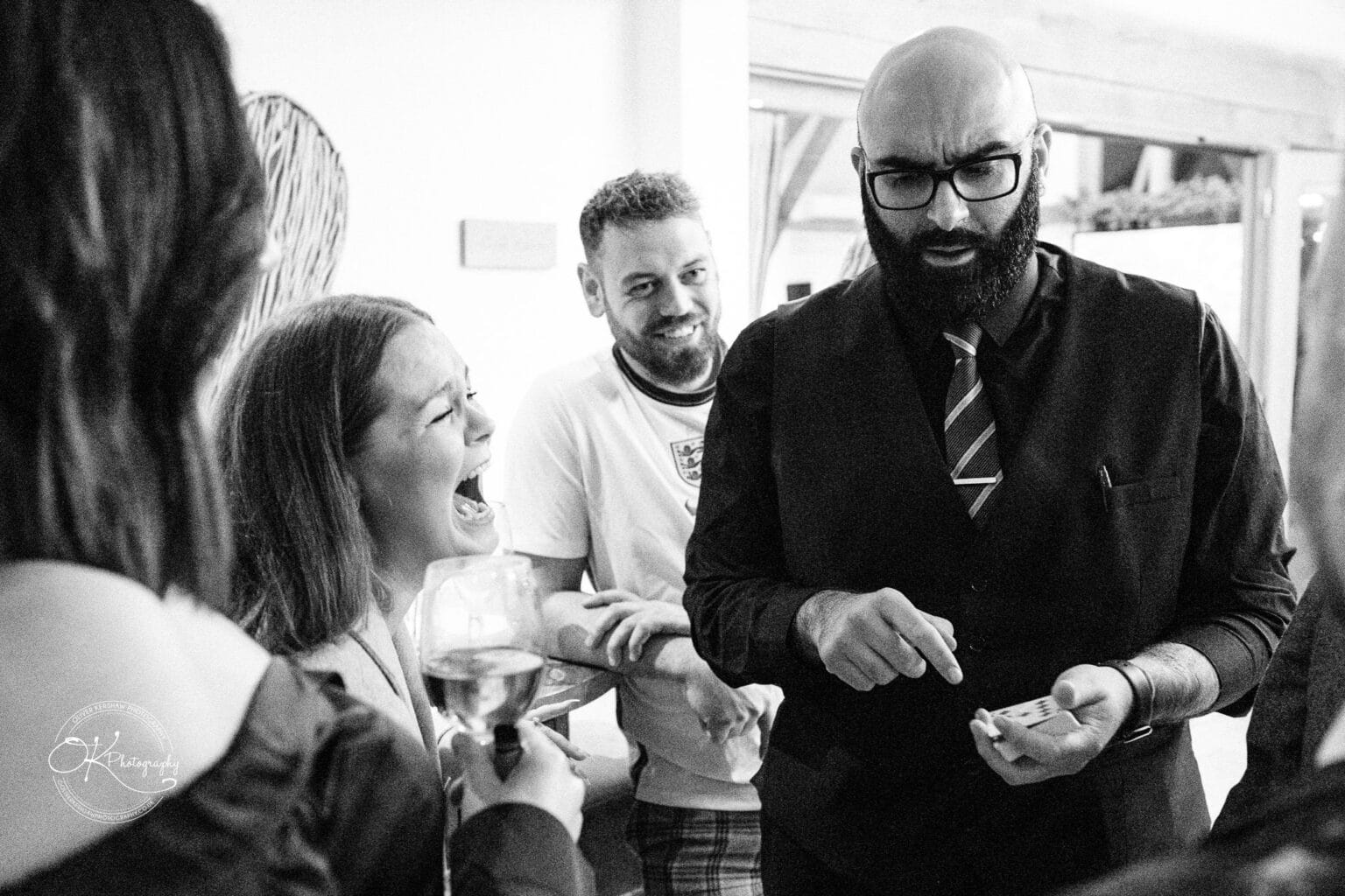 Mythe Barn Wedding photography A black and white photo of a man in a suit performing a card trick for a group of people. One woman is laughing and holding a glass of wine.