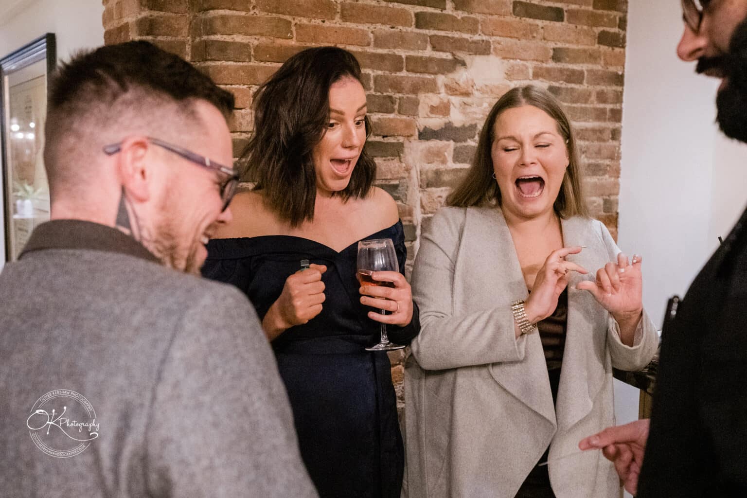 Mythe Barn Wedding photography Guests reacting with delight at a Mythe Barn wedding reception, one holding a glass of wine, as they stand in front of an exposed brick wall.