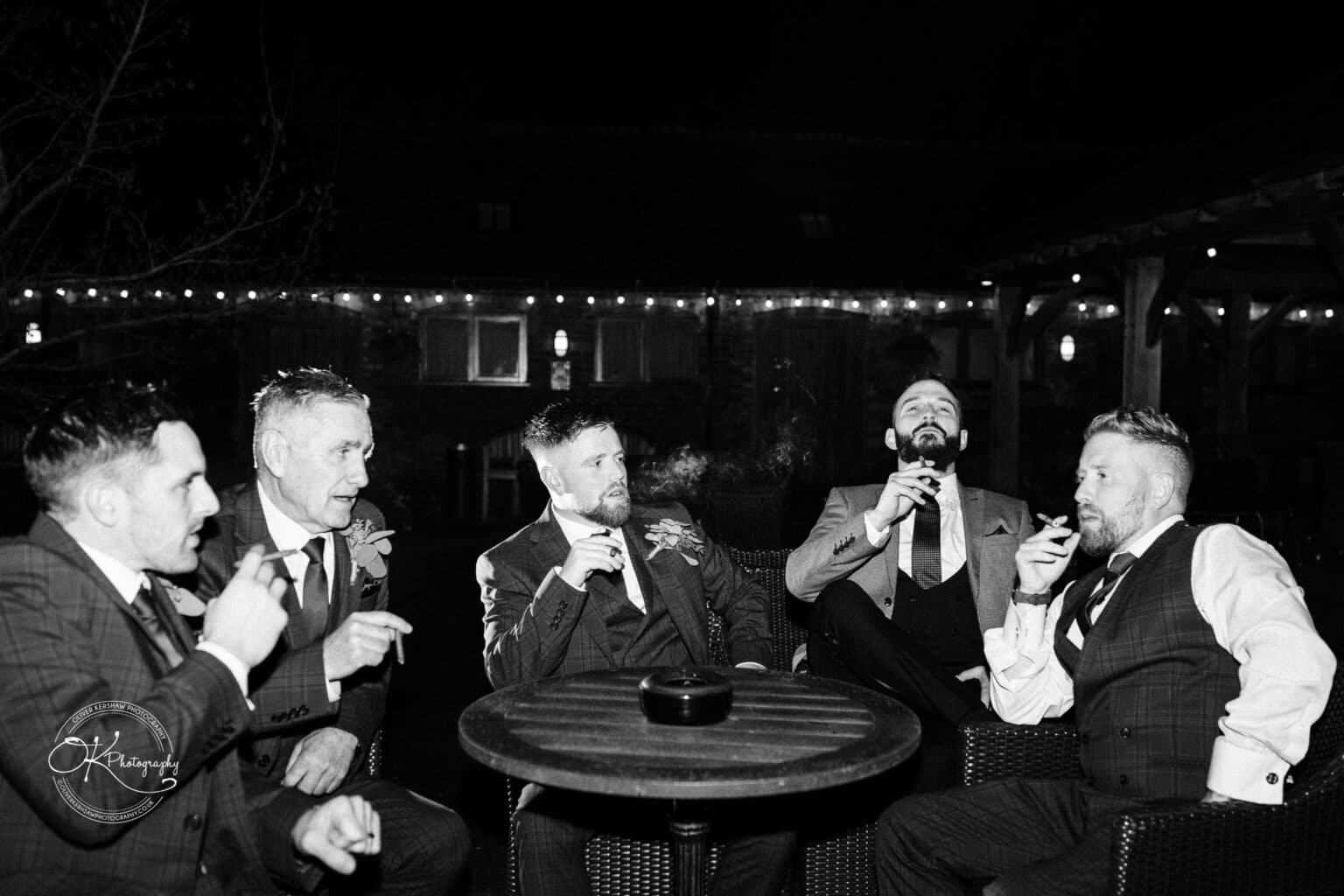 Mythe Barn Wedding photography-By-Oliver-Kershaw-Photography-OK707530 A group of five men dressed in suits, sitting around a table and smoking cigars at an outdoor seating area, with string lights in the background.