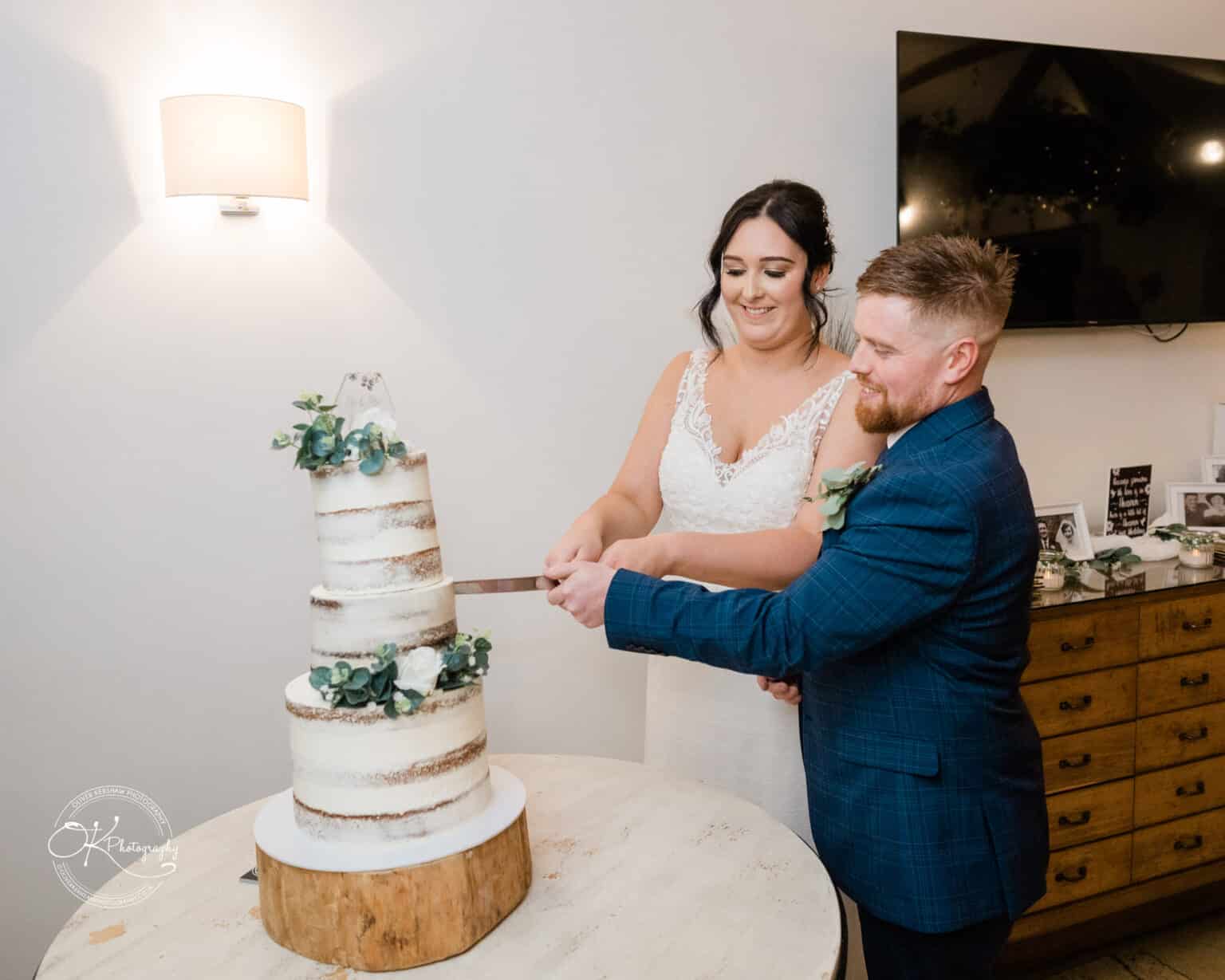 Mythe Barn Wedding photography A couple in wedding attire cutting a three-tiered naked cake decorated with greenery at a wedding reception.
