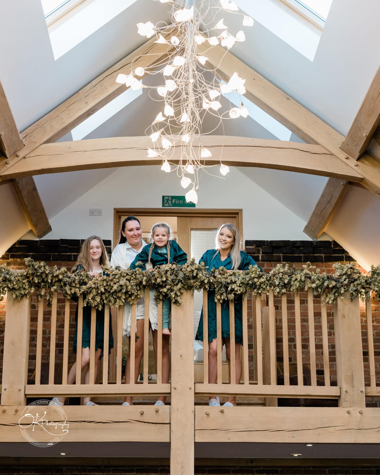 Mythe Barn Wedding photography Four women in robes standing on a decorated wooden balcony in a barn with a chandelier and exposed beams above.