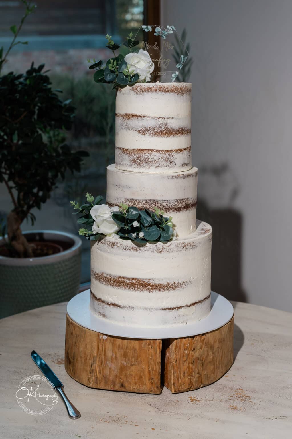 Mythe Barn Wedding photography Three-tier naked wedding cake topped with greenery and flowers, placed on a wooden stand, with a knife beside it.